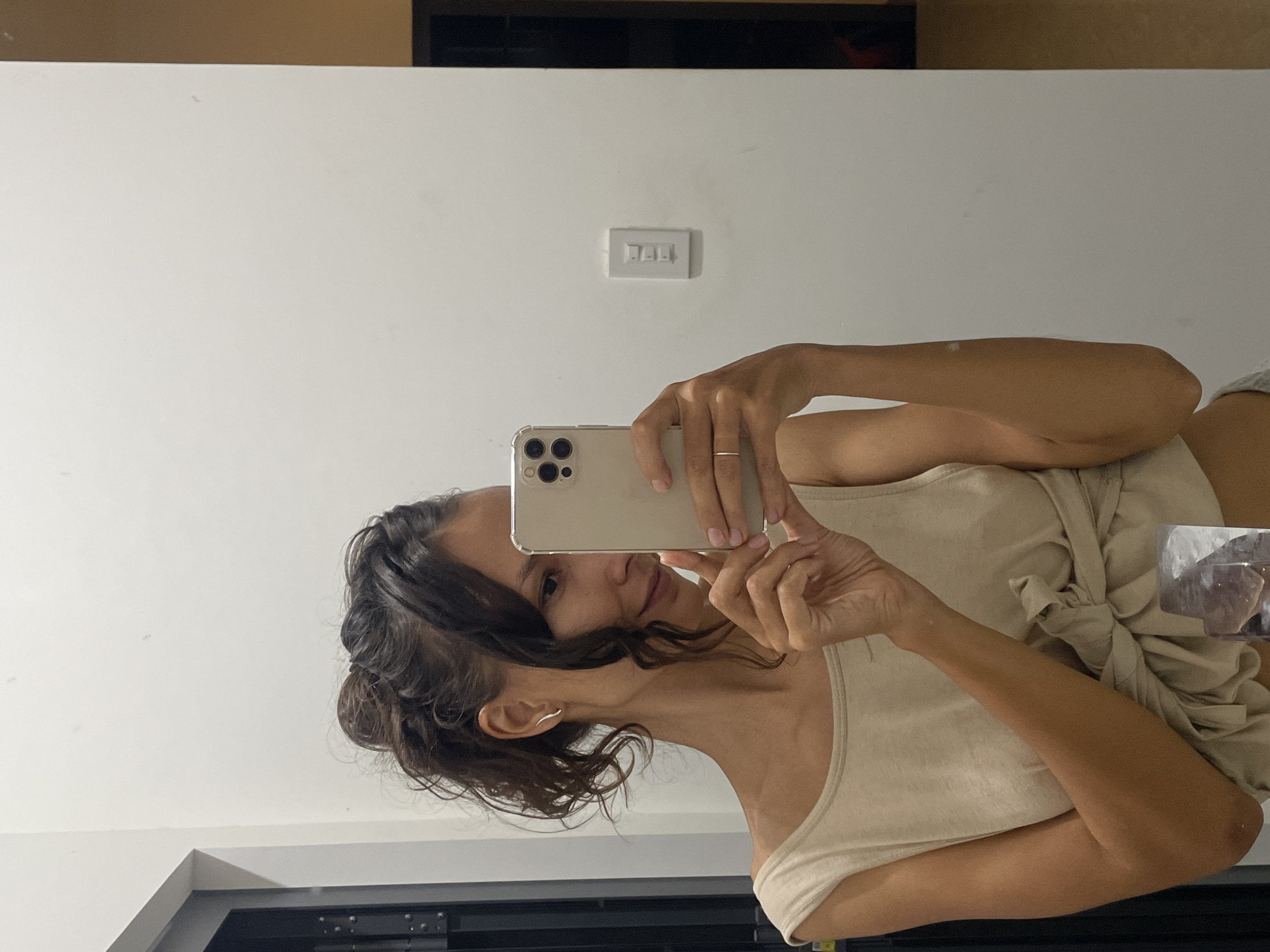 This image is a mirror selfie taken by a young woman in an indoor setting, likely a bathroom or bedroom, in Mũi Né, Vietnam. The woman is positioned on the left side of the frame, holding a light-colored smartphone with a clear case in front of her to capture her reflection. She looks towards the camera with a gentle, subtle smile.
Her dark, wavy hair is casually styled or pulled back, appearing somewhat damp, with a few strands framing her face and neck. She wears a light, possibly beige or cream-colored, off-the-shoulder tank top. A similar light-colored piece of fabric, perhaps another shirt or wrap, is tied in a knot around her waist or lower back. A thin ring is visible on the ring finger of her left hand, and she has a delicate ear cuff on her left ear.
The background behind her is a plain white wall, which is clearly reflected in the mirror. A rectangular white light switch with three toggles is mounted on this wall. To the right side of the frame, a clear glass or container holds what appears to be a decorative crystal or a large, irregularly shaped ice piece. The lighting in the room is bright and even, indicating it is daytime. No discernible text is visible in the image. The overall impression is relaxed and casual.