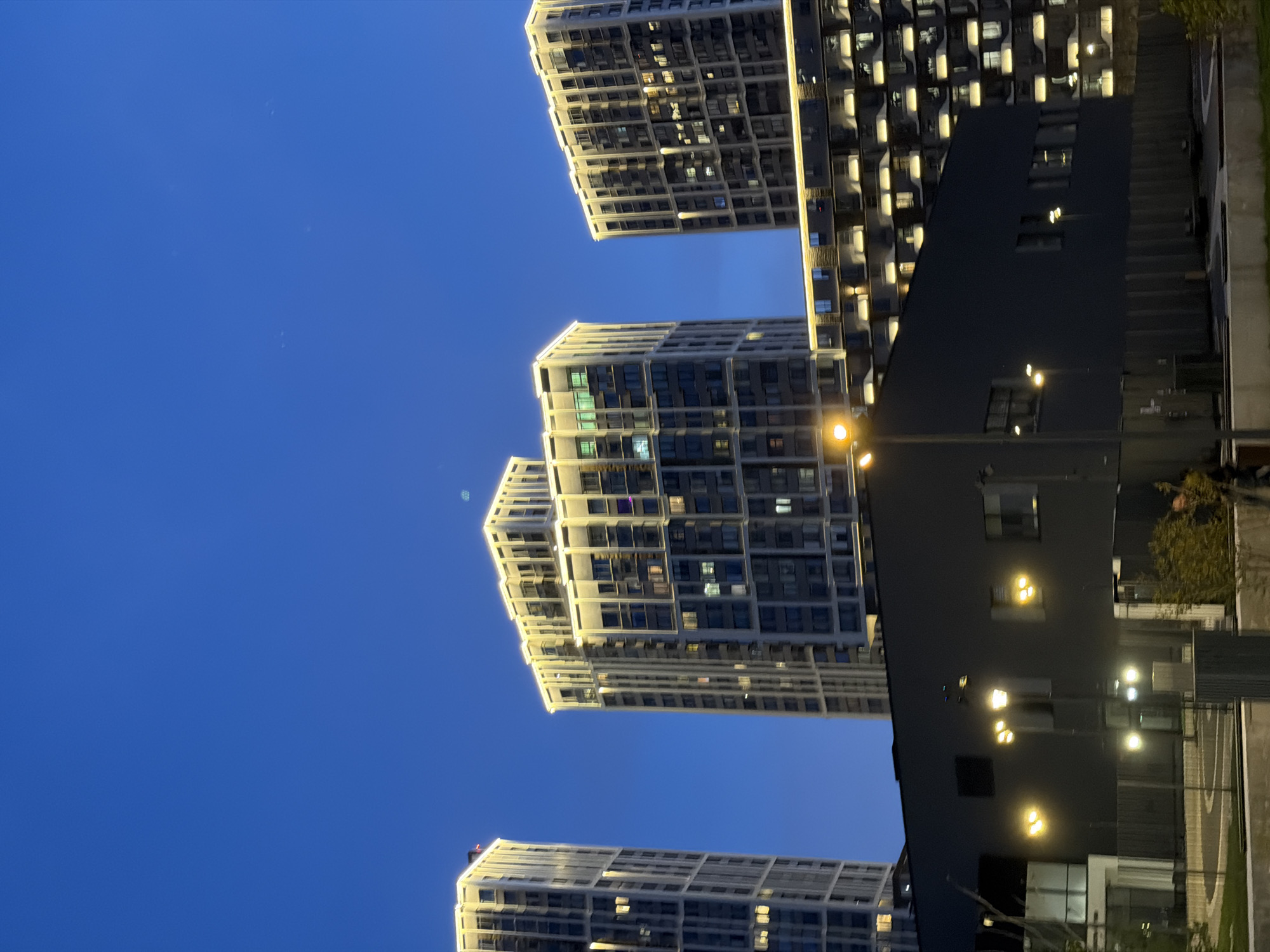 The image shows a nighttime view of modern architecture in Moscow, Russia. Three tall, illuminated residential buildings stand against a deep blue sky. The buildings have a grid-like facade with numerous windows, some lit. A darker building is on the right side of the image. Streetlights provide additional illumination, and part of a fenced area and some greenery can be seen in the lower right corner.