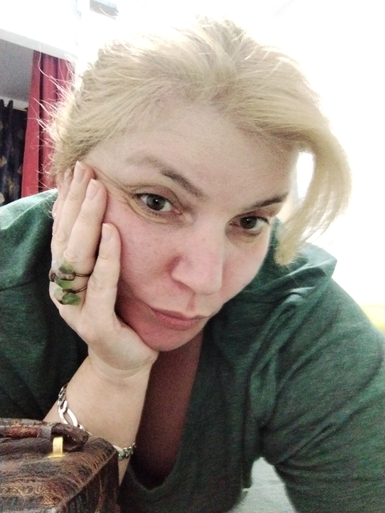 A close-up selfie shows a middle-aged Caucasian woman with blonde hair pulled back. She is wearing a green hooded sweatshirt. Her face rests on her left hand, which is adorned with two rings featuring green stones. A silver bracelet is visible on her left wrist. A dark-colored, textured box sits to the side. The background includes a partially visible curtain with dark and red tones, and a mostly white wall. There is no indication of the city of Al Mas'oudi, United Arab Emirates, or any location in the scene.