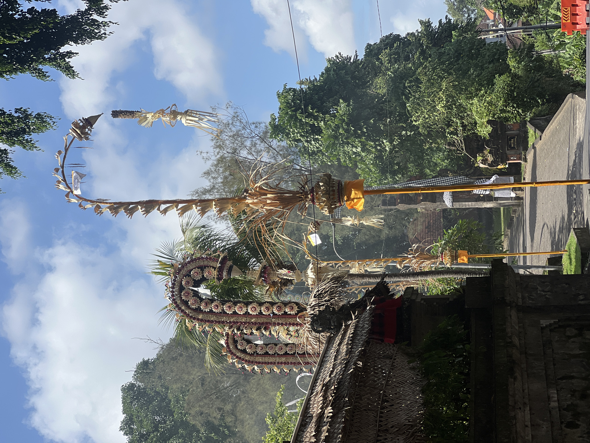 The image depicts a scene in Mengwi, Indonesia, likely related to a Balinese ceremony or festival. The primary subjects are elaborately crafted decorative structures made of natural materials like palm leaves and flowers. These are suspended from tall poles, creating an imposing and festive atmosphere. One structure, a large, serpentine form adorned with numerous small flowers and what appear to be beads, is prominently featured in the foreground, arching over a traditional Balinese building with a thatched roof. Another tall, curved decoration, resembling a lengthy, slender banner or pennant, extends upwards and to the left, crafted from dried palm fronds. In the background, lush green trees and foliage indicate a tropical environment. A paved road and parts of a stone structure, possibly a temple or gate, are visible, suggesting an outdoor setting within a community or cultural site. The sky is bright blue with scattered white clouds, indicating daytime. There is no visible text in the image. The overall impression is one of vibrant cultural expression and natural beauty.