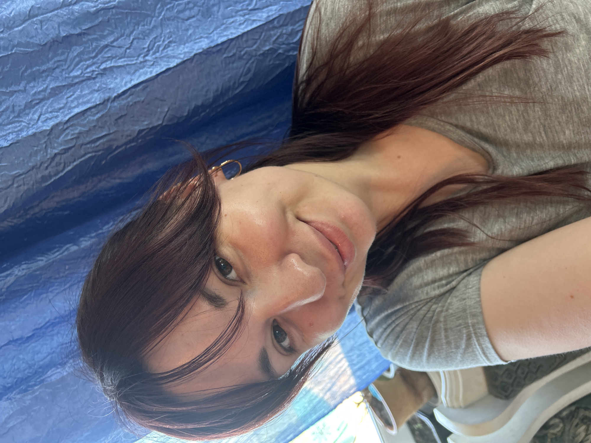 A young woman with dark hair and brown eyes is captured in a close-up, selfie-style portrait. She is wearing a grey t-shirt and a golden hoop earring in her left ear. The background consists of a wrinkled blue fabric, possibly a curtain or drape, and a hint of a window with sunlight streaming through. The scene appears to be indoors, with a potted plant visible on a ledge to the left. The overall lighting is bright and natural. The location of this image is in the city of Fokino, Russia.