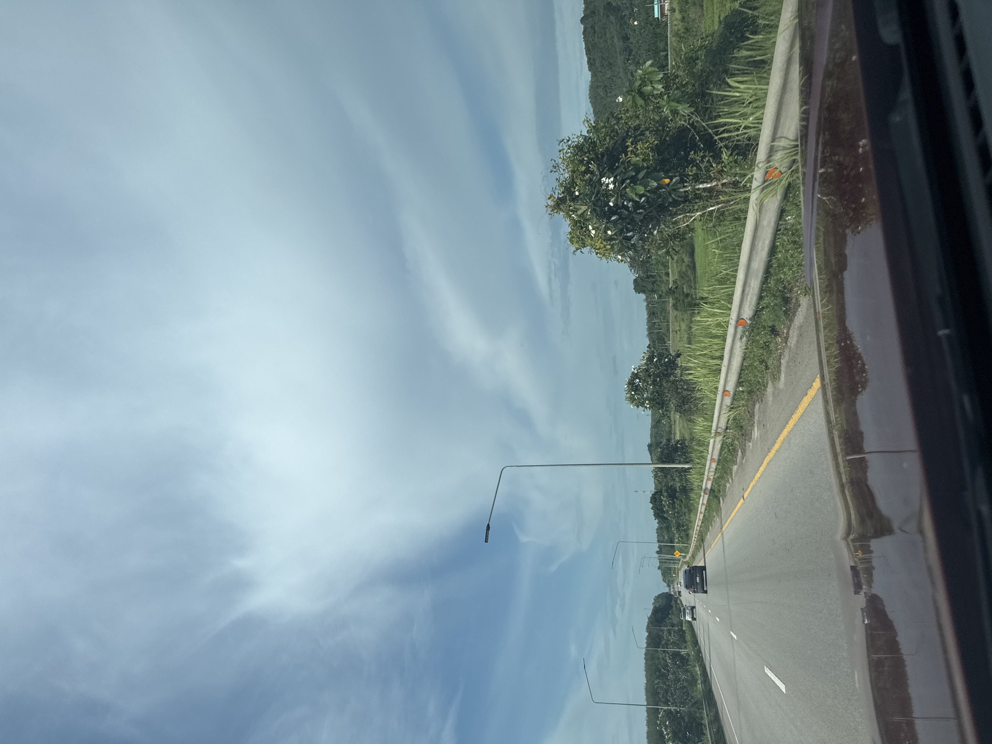The image is a photograph taken from the perspective of someone driving on a highway in Tambon Khao Tok, Thailand. The highway has multiple lanes and is bordered by a guardrail and lush green vegetation, including trees and tall grass. In the distance, rolling hills covered in trees are visible. The sky is a prominent feature, with dramatic, wispy white clouds against a blue backdrop. Several streetlights line the side of the road. Cars are visible on the highway, indicating it is in use. The foreground shows the hood of the vehicle from which the photo is taken, reflecting the sky and surrounding landscape. The overall impression is that of a scenic drive through a rural or semi-rural area.