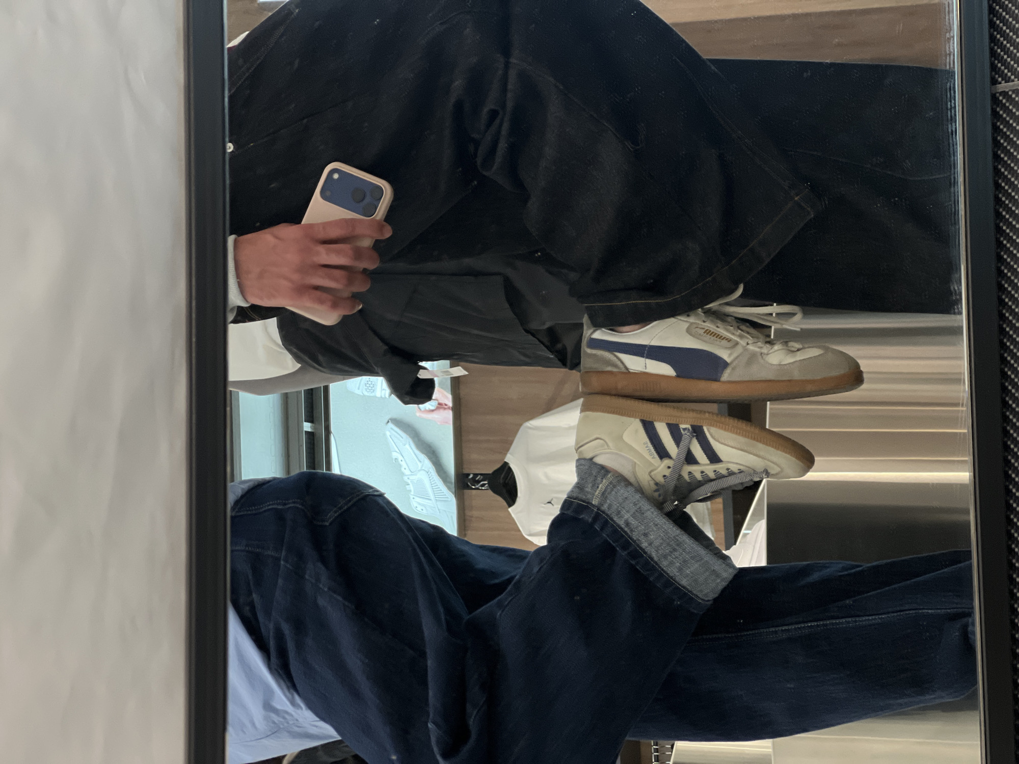 The image shows a mirror reflection of a person taking a selfie. The person is wearing dark-colored jeans, a white long-sleeved shirt, and two different sneakers: a Puma sneaker on the top and an Adidas sneaker on the bottom. The person is holding a phone with a light pink case. The background is blurred, suggesting an indoor environment. There is no information in the image to indicate this picture was taken in Yerevan, Armenia.