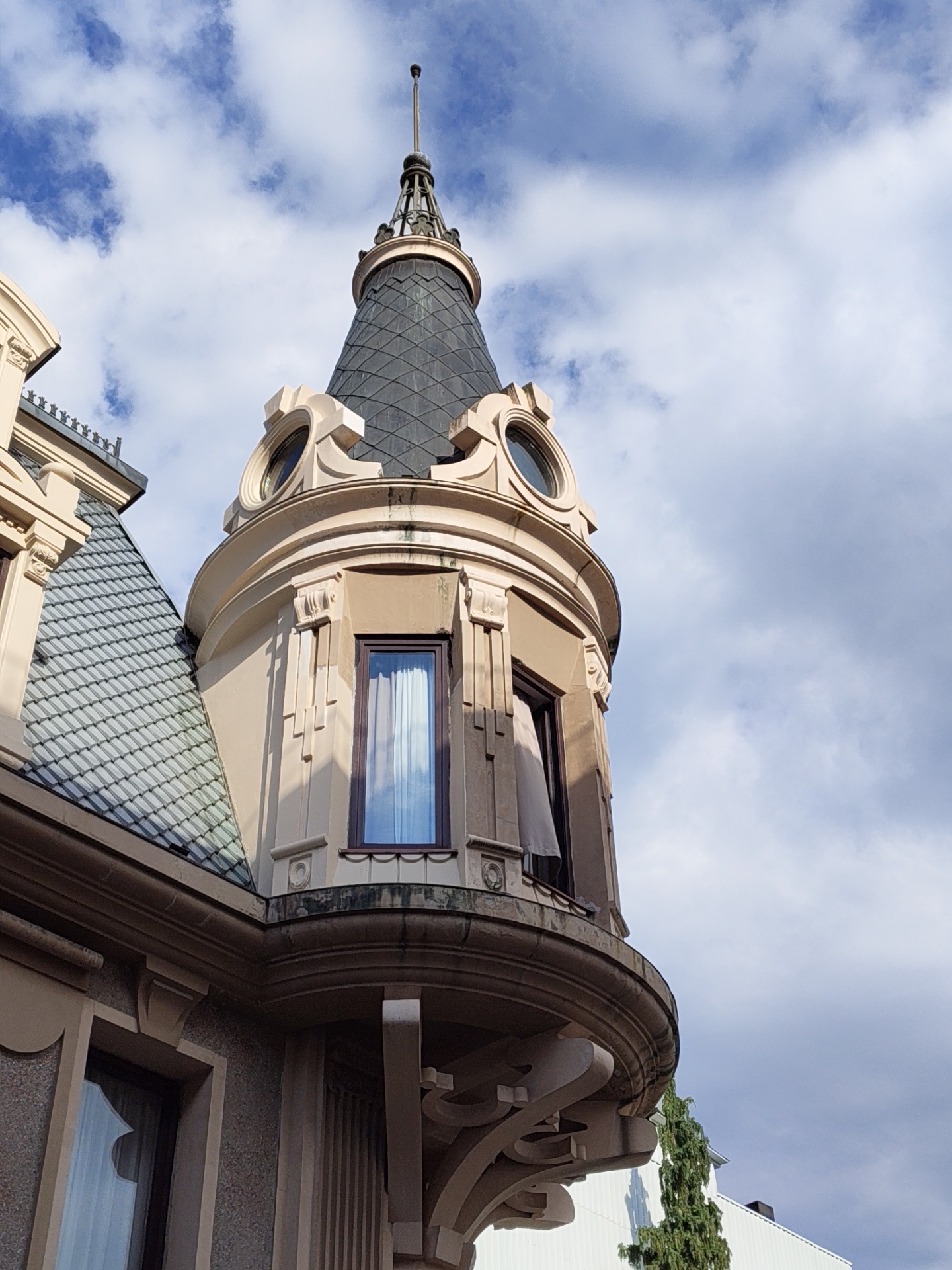 The image shows a close-up, low-angle view of a turret on a building, with a cloudy blue sky in the background. The turret has a conical, dark-tiled roof topped with a spire. The turret's body is a pale peachy color, with decorative architectural elements, including a round window at the top and a rectangular window below. A curved balcony juts out from the turret. To the left, part of another section of the building is visible, with a steeply pitched, grey-tiled roof and decorative eaves. The architectural style appears to be somewhat ornate, possibly late 19th or early 20th century. No people are visible in the image. The scene is likely from a street in Batumi, Georgia, given the architectural style and the presence of a tall, slender evergreen tree visible in the lower right portion of the frame, which is common in coastal regions like Batumi. The overall impression is of a historic building bathed in natural light under a dynamic sky.