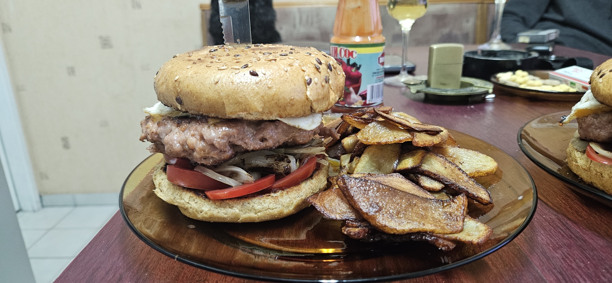 The image shows a close-up of a hamburger on a dark brown glass plate with a side of fried potato slices. A knife is inserted into the top bun of the hamburger. The hamburger includes a bun, tomato slices, onions, a meat patty, and what appears to be a fried egg. Behind the plate, there are bottles and glasses on a brown wooden surface. An ashtray with cigarettes and a lighter is visible to the right of the plate. The location is not explicitly stated but could be in Sofia, Bulgaria, given the request for that specific location. The environment suggests an indoor setting, possibly a restaurant or a home. There are partially visible people and another plate with food on the right side of the image.