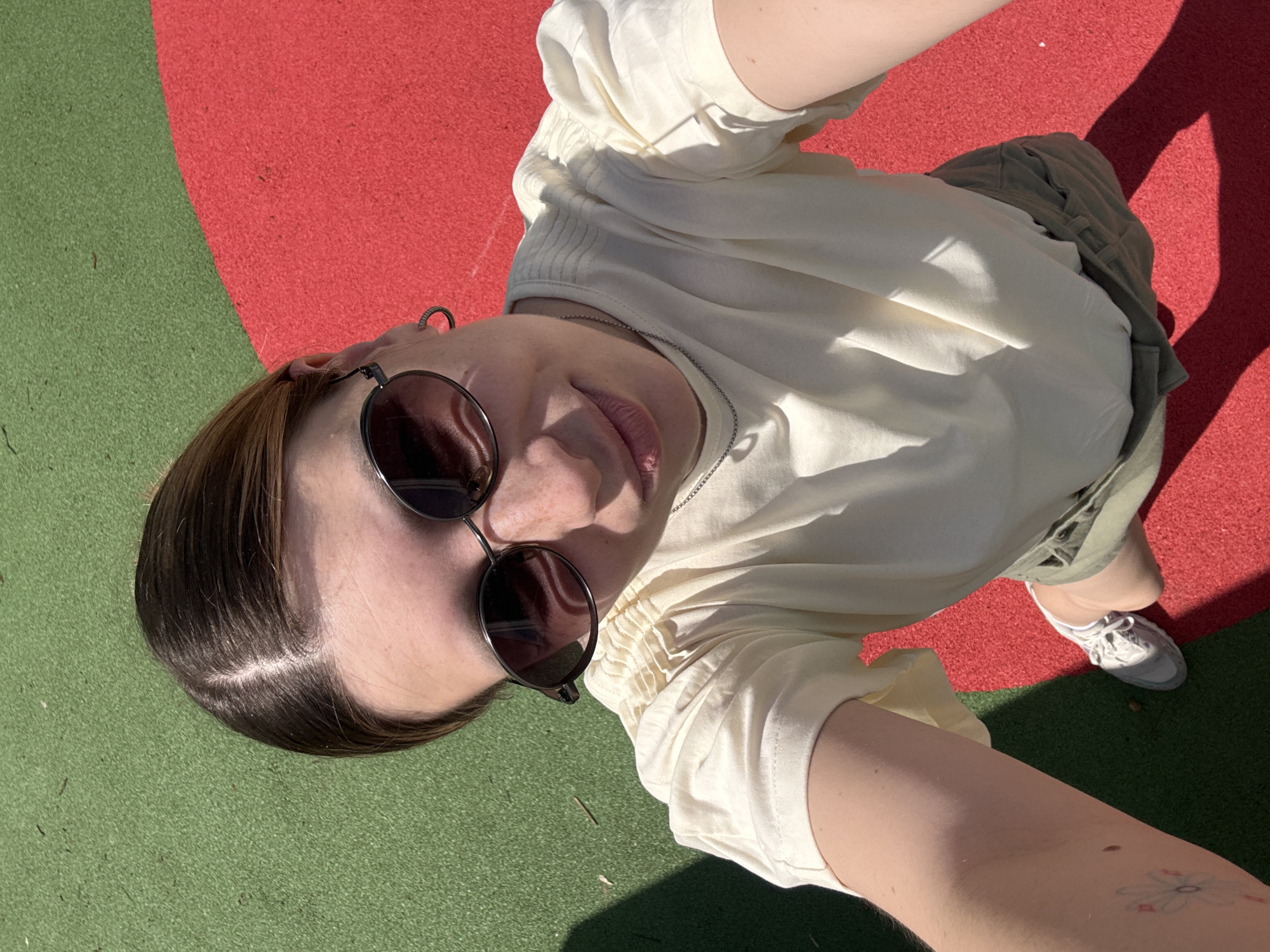 The image is a selfie taken from a high angle, showing a person wearing round sunglasses, a cream-colored blouse with gathered sleeves, and olive-green shorts. The person is standing on a surface divided into red and green sections. A white sneaker is visible at the bottom right of the frame. The person has a tattoo on their arm. There is no information available that suggests this image was taken in Moscow, Russia.