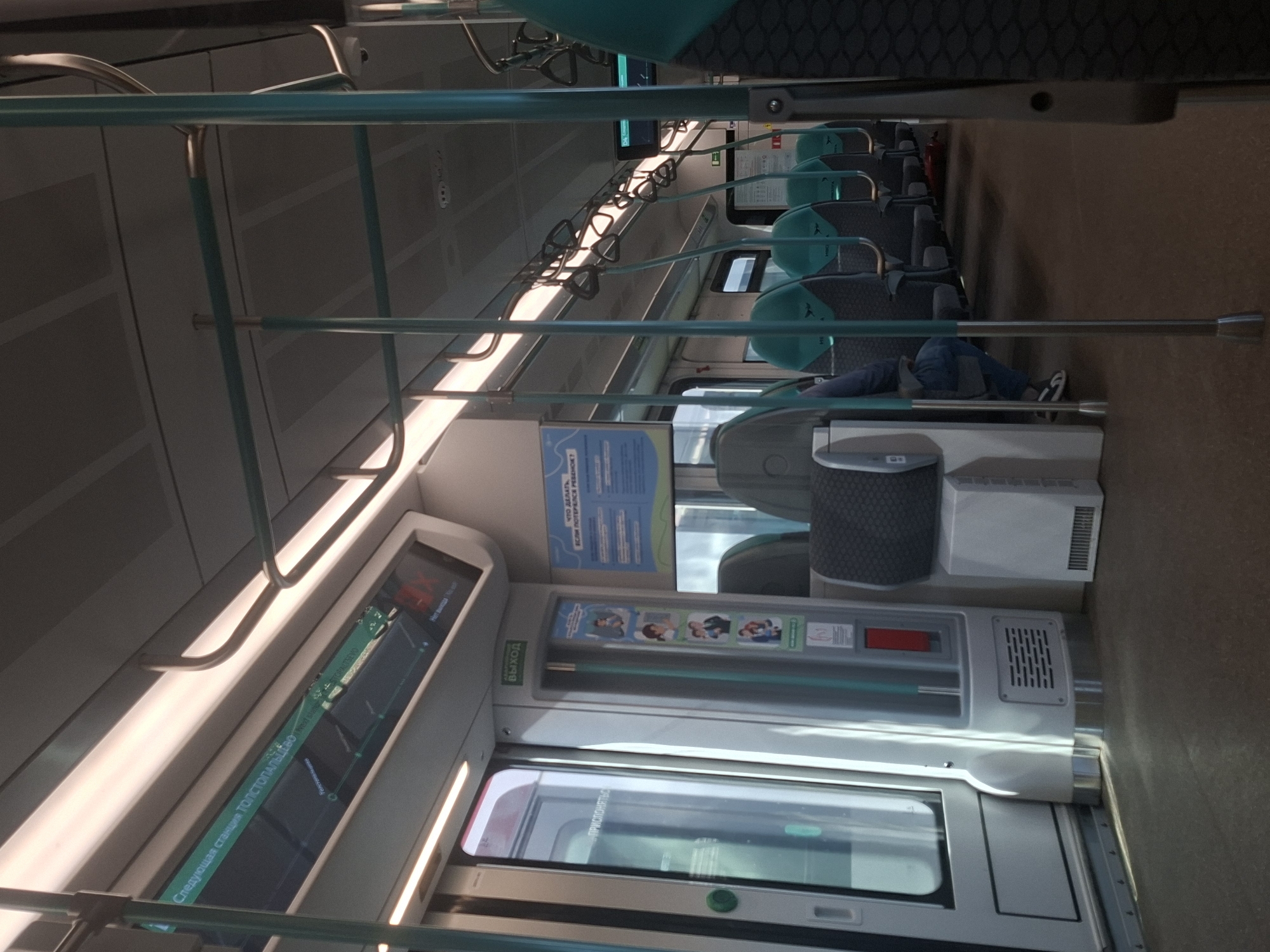 The image depicts the interior of a public transport vehicle, likely a train or metro car, with an emphasis on the seating area and internal structures. The color palette is dominated by cool tones, including silver, grey, and teal, with accents of white and red. Fluorescent lighting illuminates the scene from above, casting a soft glow.
In the foreground, to the right, a section of a dark, textured seat is visible. Moving towards the center, rows of teal and grey seats with chrome accents are arranged, some occupied and some empty. A person wearing jeans and a blue jacket is seated on the left side, facing away from the camera. Several vertical teal poles and horizontal metal bars are strategically placed throughout the carriage, serving as handrails and structural supports.
On the left side of the image, a silver door with a large glass panel is present. Above the door, a digital display shows information in Russian, indicating "Следующая станция ТОЛСТОПАЛЬЦЕВО" which translates to "Next station TOLSTOPALTSEVO." Another sign with Russian text, "ВЫХОД" (Exit), is positioned above a panel containing small, illustrative graphics.
Further down the carriage, more rows of seats are visible, with a dark floor covering the ground. The ceiling is comprised of rectangular panels with integrated lighting. The overall impression is of a clean and functional interior of a modern commuter vehicle. The mention of "TOLSTOPALTSEVO" suggests a location within or near Moscow, Russia, given its proximity to Vnukovo International Airport.