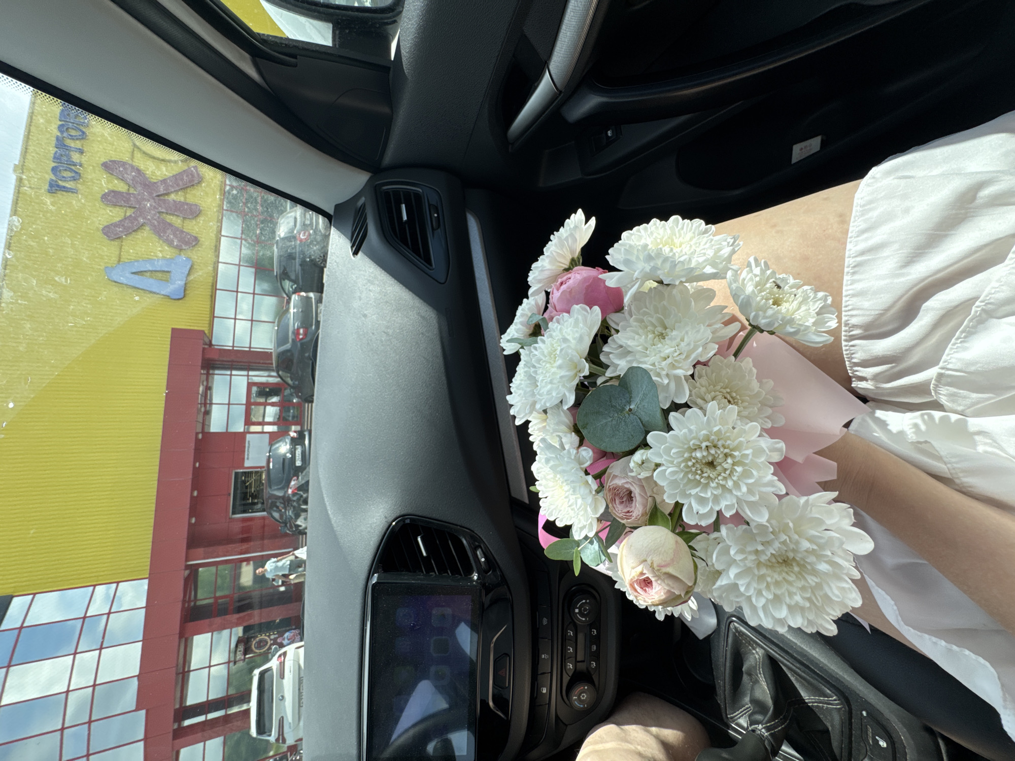 The image shows the interior of a car, viewed from the passenger seat looking towards the dashboard and out the windshield. A bouquet of white chrysanthemums and pink roses is held in the foreground, resting on the lap of an unseen person. The dashboard features a large touchscreen display and climate controls. Outside the car, a yellow building with red trim is visible, identified by the text "Торговый Дом" (Department Store) and the Cyrillic letters "ДЖ" in blue and pink. Several cars are parked in front of the building. The scene suggests a commercial area in Chaikovsky, Russia, on a sunny day.