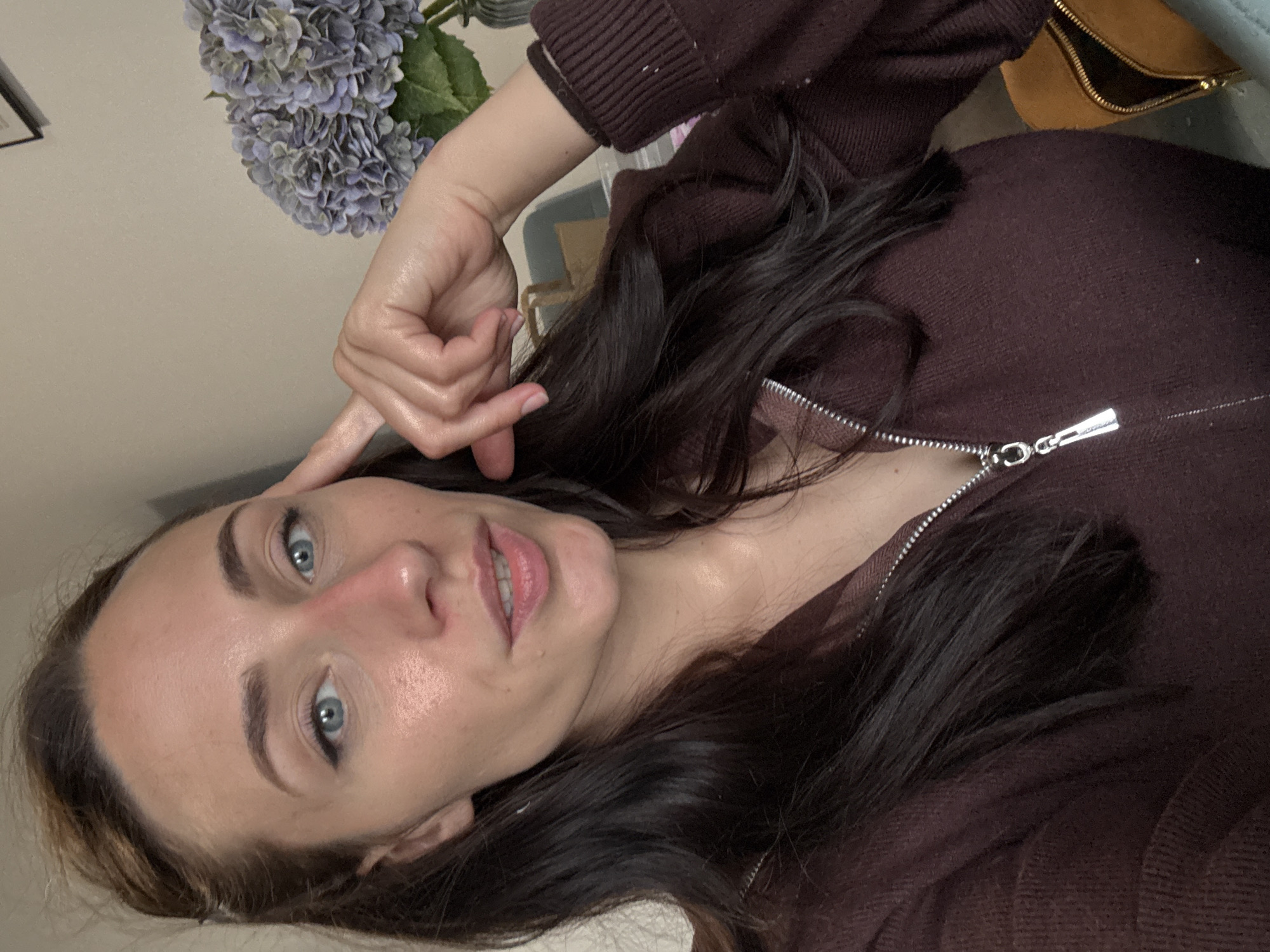 A woman with blue eyes and long dark hair is depicted from a close-up, angled perspective. She is wearing a brown zip-up sweater. Her left hand is raised, with her index finger touching her forehead. A bouquet of blue hydrangeas is partially visible in the background. The background is a plain wall.