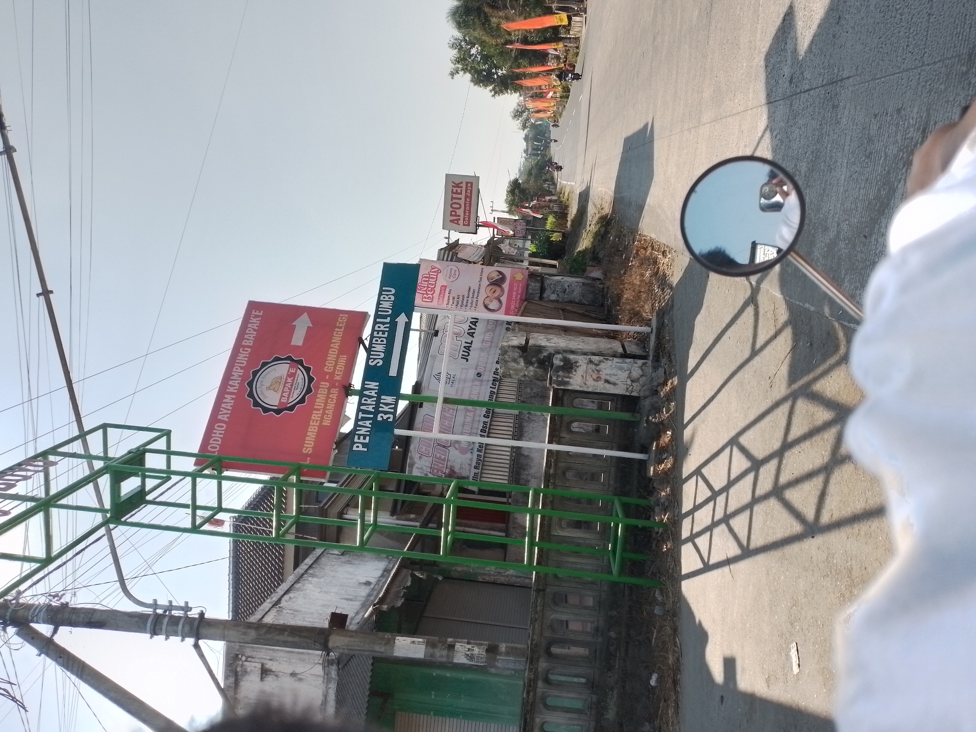 The image depicts a street scene in Ngancar, Indonesia, likely during the day. A green metal frame stands near a concrete fence with arched openings. A red billboard advertises "LODHO AYAM KAMPUNG BAPAK'E" with an arrow pointing right, listing "SUMBERLUMBU - GONDANGLEGI - NGANCAR - KEDIRI." Below this, a blue sign reads "PENATARAN 3KM SUMBER LOMBO". To the right of the green frame, other signs are visible, including one for "Kim Beauty" offering various treatments, and an "APOTEK COLORANTE JAYA". The road stretches ahead, lined with orange flags and trees. Several motorcycles are present on the road, with riders visible on some. A motorcycle side mirror in the foreground reflects the sky and a person, presumably the photographer, holding a phone. The ground is paved asphalt, with shadows cast by the green frame and power lines overhead. The overall environment suggests a rural or semi-urban setting with commercial establishments.
