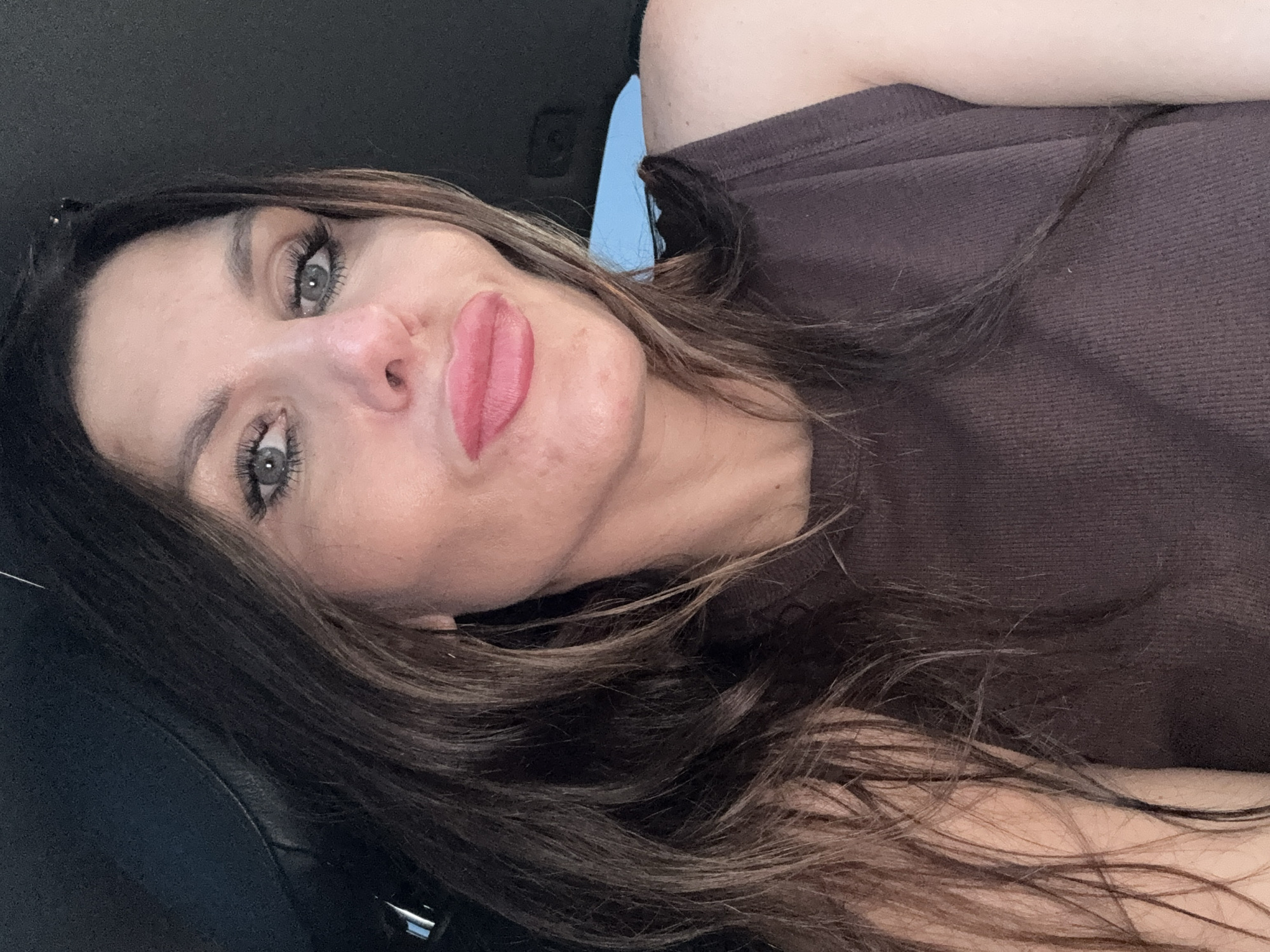 A woman is sitting in the driver's seat of a car, taking a selfie. She has long, dark brown hair and is wearing a brown, ribbed tank top. Her makeup is visible, including mascara and lipstick. The background of the image shows the interior of a car, with a dark headliner and a sliver of blue sky visible through the windshield. There is no visible indication of the city of Chaikovsky, Russia, or any specific location within it.