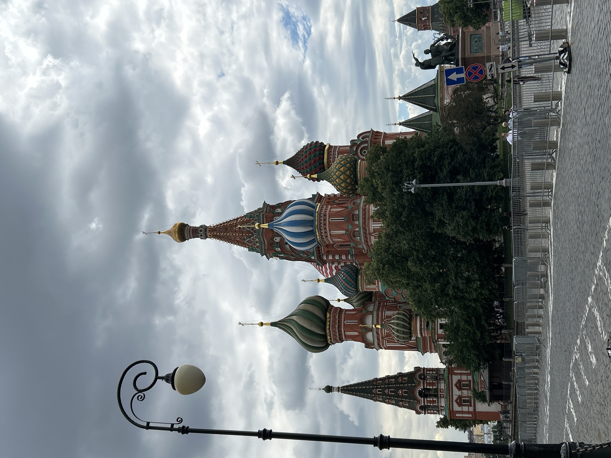The image depicts Saint Basil's Cathedral in Moscow, Russia, on a cloudy day. The iconic cathedral, with its vibrant red walls and multi-colored onion domes, dominates the background. In the foreground, a lamppost with a decorative curved arm and an opaque globe is visible on the left. A paved cobblestone street with white markings runs across the bottom of the frame. Metal barriers are set up along the street, with a few people standing behind them. A statue of two figures is visible in the middle ground to the right of the cathedral, accompanied by traffic signs. Lush green trees are interspersed around the base of the cathedral.