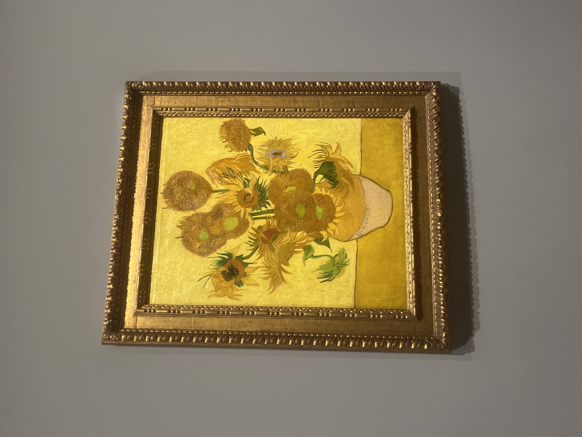 A framed oil painting of sunflowers hangs on a grey wall. The painting features a vase of sunflowers against a yellow backdrop, rendered in the distinct style of Vincent van Gogh. The frame is ornate, gold-colored, and appears to be made of wood. The location is likely the Van Gogh Museum in Amsterdam, Netherlands.