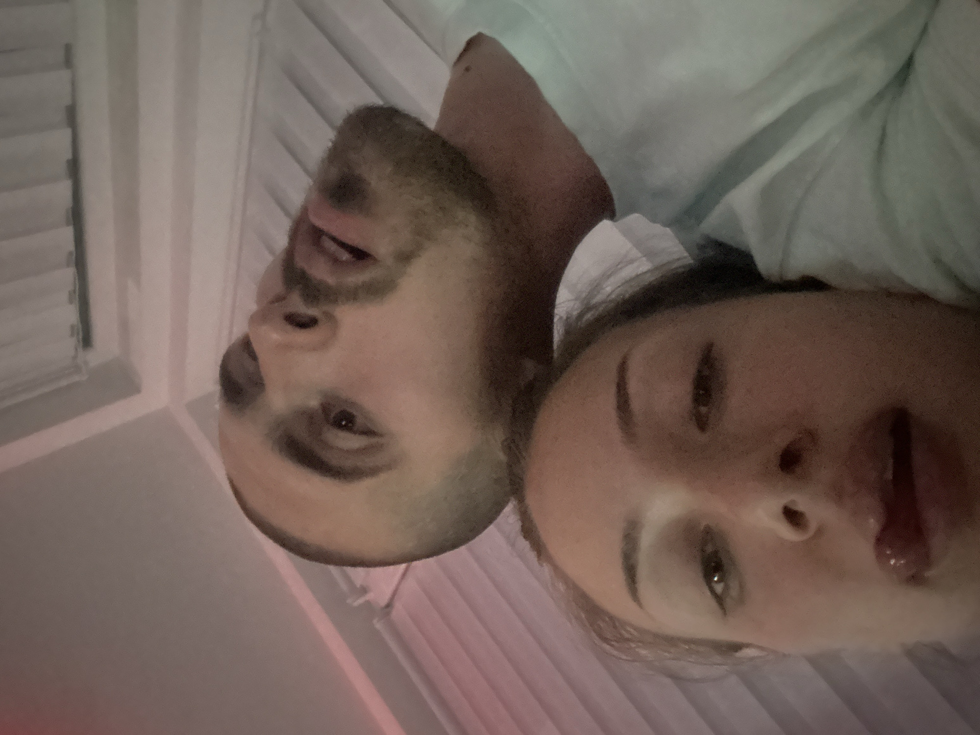 Two people are lying down next to each other. The person on the left has short hair, a beard, and is looking towards the camera. The person on the right has long hair and is also looking towards the camera with their mouth open. They appear to be lying on a bed, in a room with closed blinds. The location is unknown.