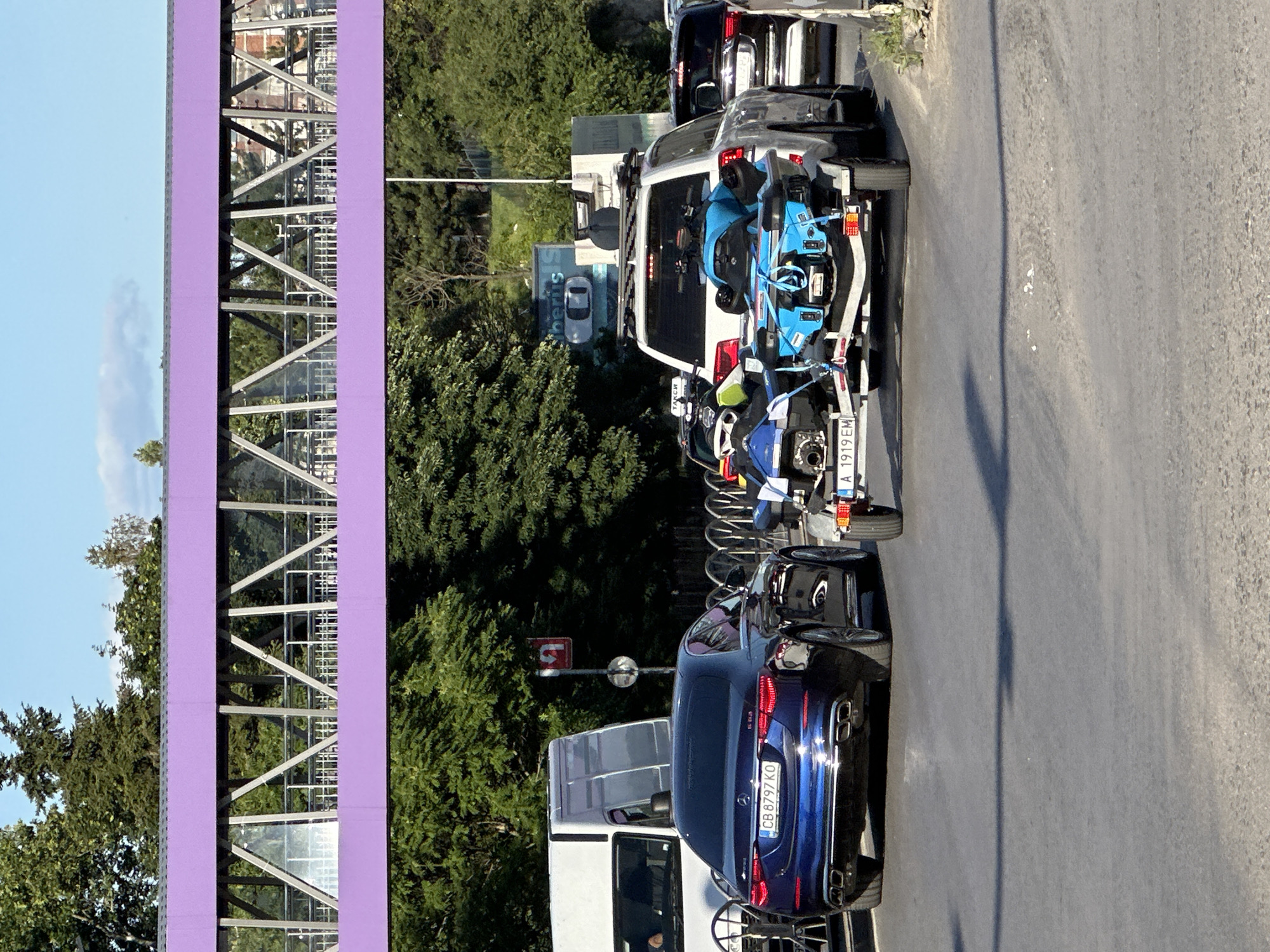 This image captures a street scene in Veliko Tarnovo, Bulgaria. A dark blue Mercedes-Benz sedan with license plate CB 8797 KO is in the foreground, facing away from the camera. It is in traffic, along with several other vehicles. Directly behind the Mercedes is a white SUV towing a trailer with two jet skis. The trailer has a license plate beginning with A 1919 EM. Beyond the vehicles, a pedestrian bridge with a purple railing and a metal structure can be seen, spanning over the road. Lush green trees line the sides of the road and a bright blue sky with a few clouds is visible above. The overall impression is of a sunny day with moderate traffic on an urban road.