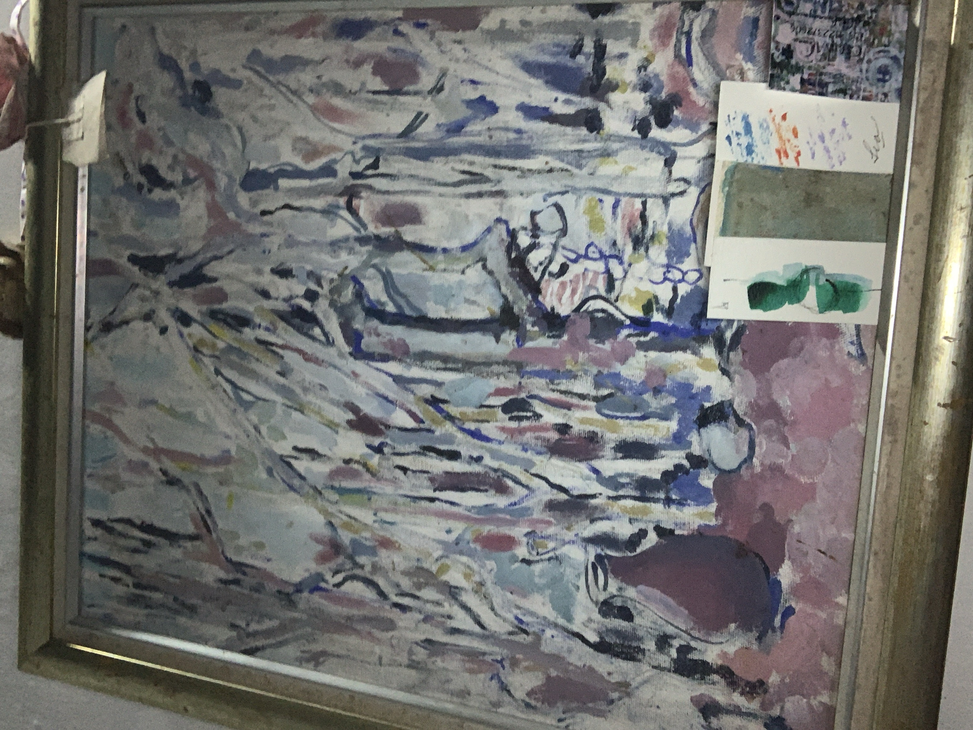 A framed abstract painting is displayed. The painting features broad brushstrokes in shades of white, blue, pink, and purple, creating a somewhat chaotic yet harmonious composition. In the foreground, at the bottom right, a small collection of cards or postcards are placed. One card has three distinct sections: a green abstract figure on the left, a textured gray rectangle in the middle, and a section with blue and orange abstract marks above purple smudges on the right, with a handwritten signature "Leo" below it. To the right of this, another small card with a complex pattern is visible. The painting is housed in a metallic gold-colored frame. The backdrop appears to be a wall. The scene is indoors. No specific location in Tacarigua, Venezuela, can be determined from the image.