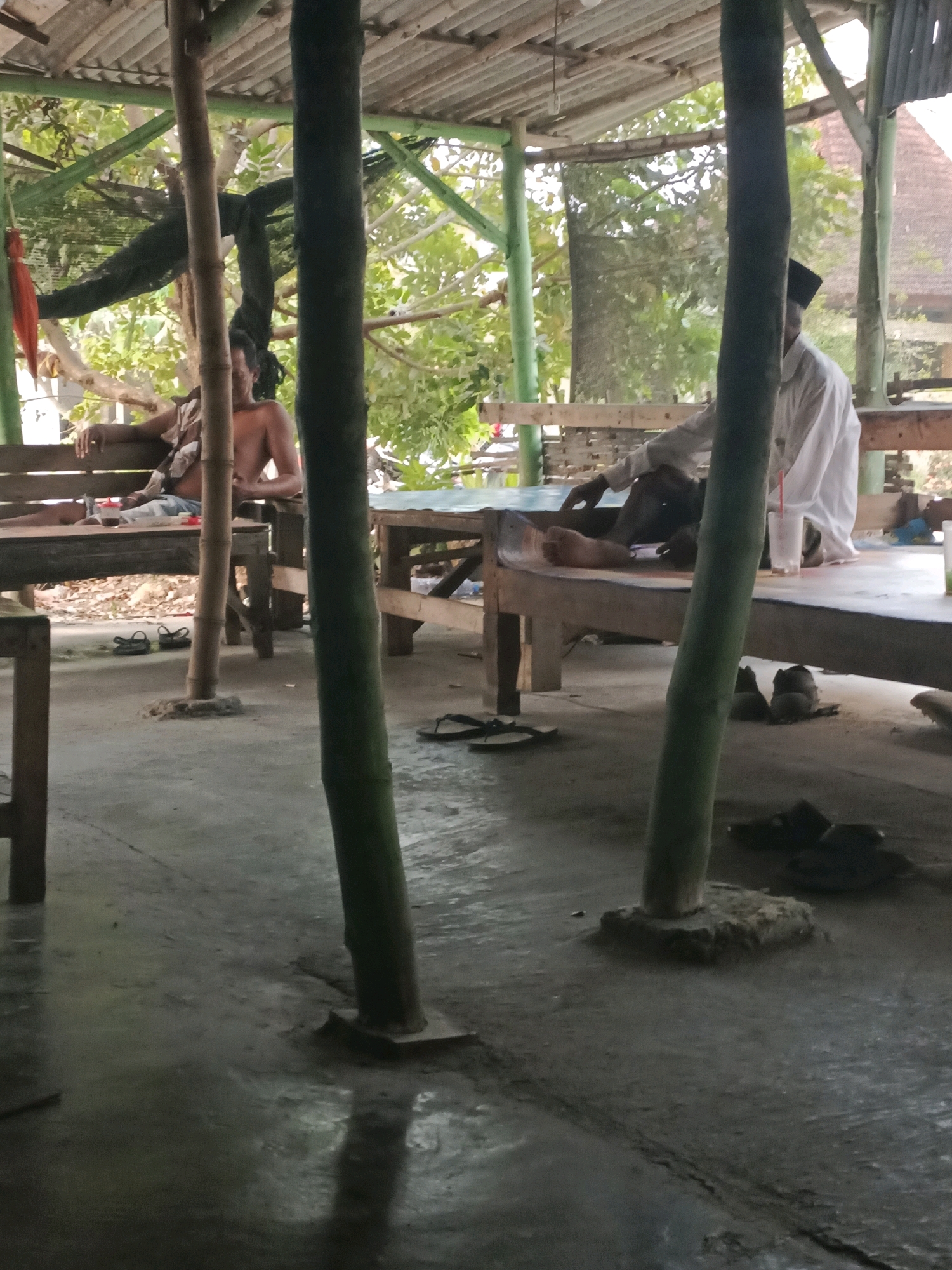 The image shows a scene at an open-air establishment in Rancang Kencono, Indonesia, likely a roadside eatery or resting spot. The structure has a corrugated metal roof supported by bamboo poles and wooden beams. Two men are visible. In the foreground, a shirtless man sits on a wooden bench, draped with fabric over his shoulders. He appears to be a customer. Further back, a man wearing a white shirt and a black peci (cap) is seated at a wooden table, also appearing to be a customer. On the table, there are glasses and a plastic cup with a red straw. The ground is concrete, with some debris and a few pairs of sandals scattered around. Trees and foliage are visible in the background, suggesting a tropical or subtropical environment. The lighting indicates it is daytime, with sunlight filtering through the canopy.