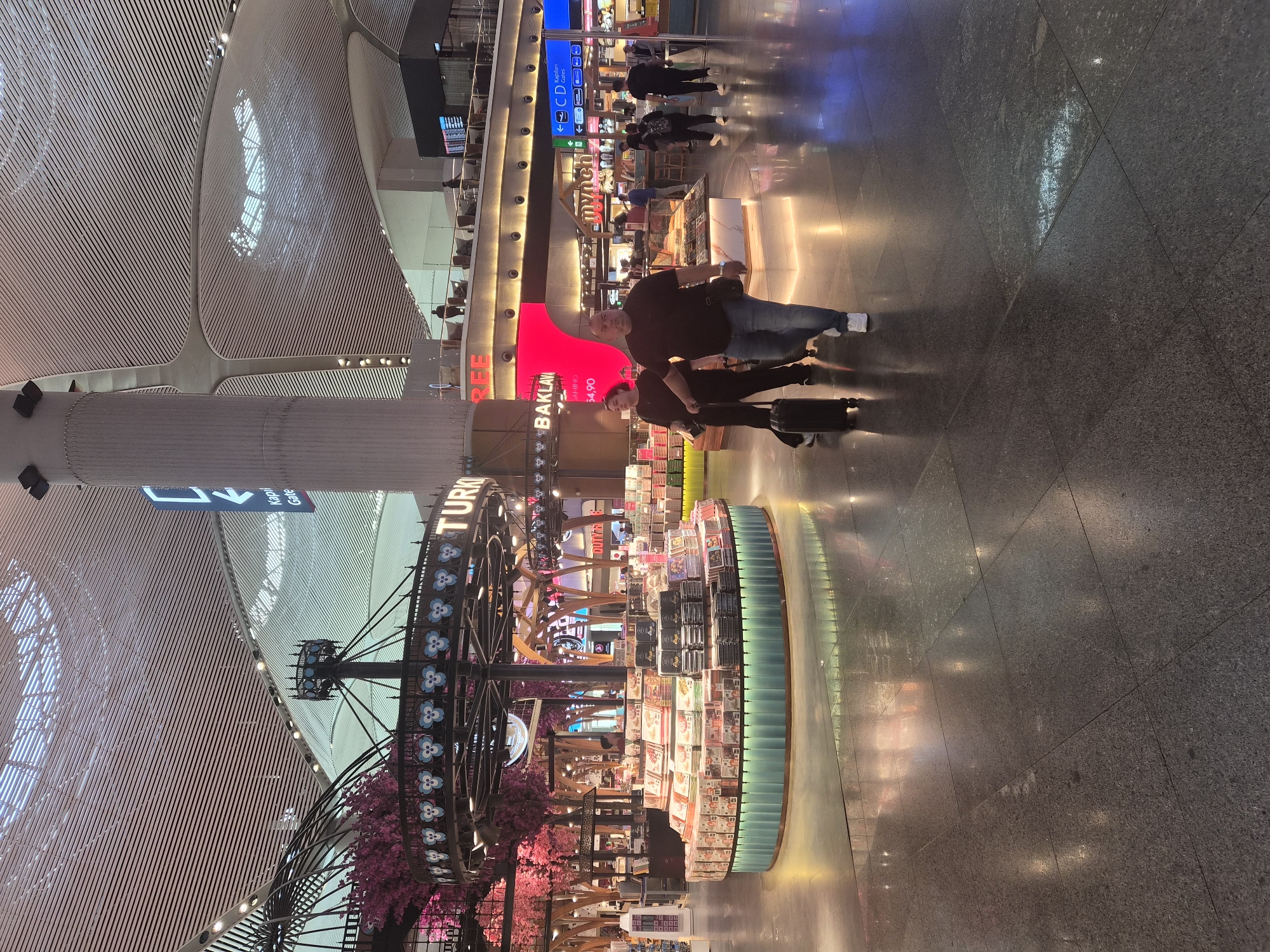 This is a full shot of the interior of an airport terminal in Arnavutköy, Turkey. The scene is dominated by a modern, high-ceilinged space with a slatted white ceiling structure. In the foreground, a couple walks away from the camera towards the left. The man is in the center, wearing a dark t-shirt and blue jeans, and is pulling a black suitcase. To his left, a woman in a dark t-shirt and pants walks beside him, also pulling a suitcase.
In the mid-ground, a circular display stand is filled with boxes of Turkish delight and baklava. Behind this, a neon sign reads "TURKISH" and "BAKLAVA". Further back, there are various shops and seating areas visible. People can be seen walking and sitting in these areas. Blue directional signs indicate "Kapı / Gate". The floor is polished dark stone, reflecting the overhead lights and the displays. The overall atmosphere is busy and commercial, typical of a large international airport.