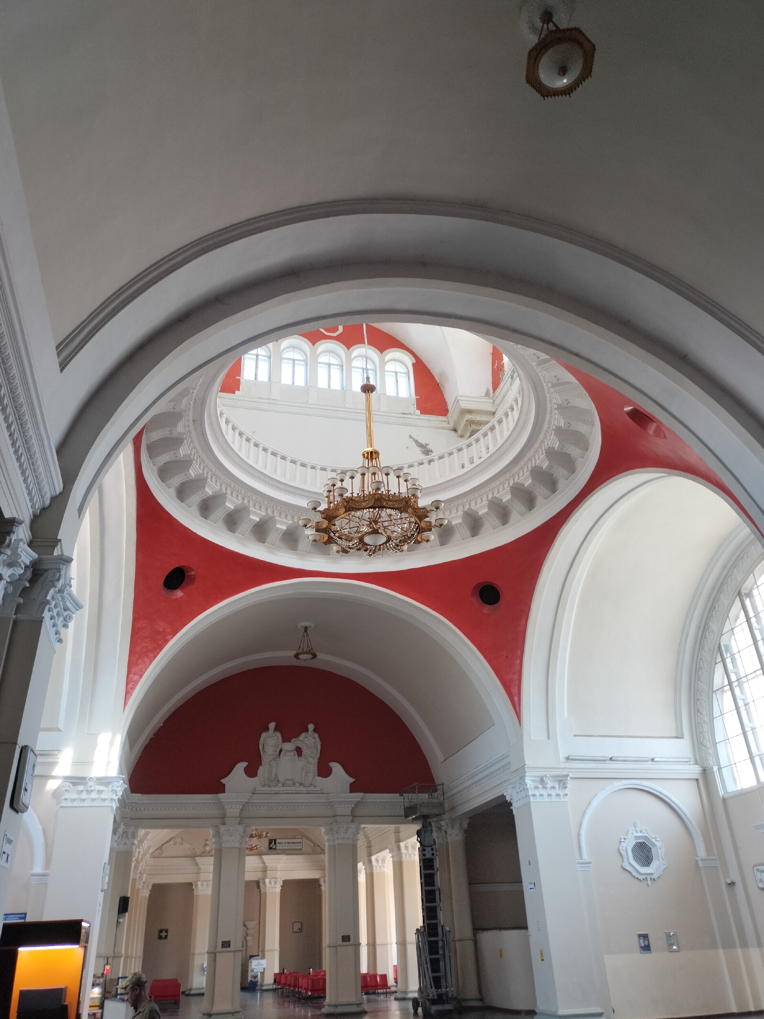 The image shows the interior of the Chernihiv railway station in Chernihiv, Ukraine. The architecture is grand, with high vaulted ceilings and elaborate detailing. A large, ornate chandelier hangs prominently in the center of the frame, illuminated by natural light streaming in from large windows. The walls are painted in a combination of white and vibrant red, adding a striking visual contrast. In the background, there are rows of seating and architectural columns, suggesting a waiting area or hall. The scene appears calm and empty, with no significant activity or people clearly visible.