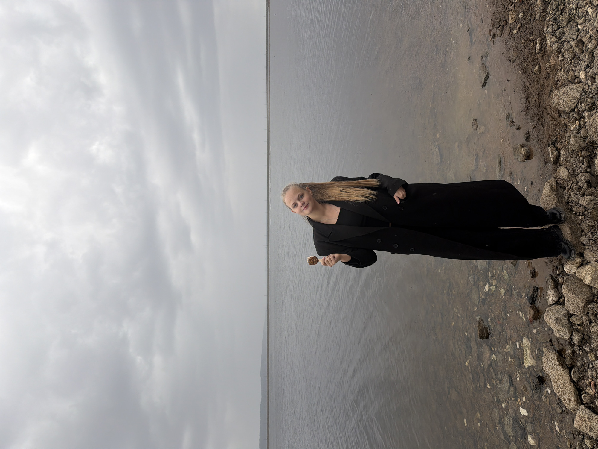 A vertical, wide shot captures a young woman standing on a rocky shore, eating an ice cream cone, against a backdrop of a grey, cloudy sky and a body of water. The woman, positioned to the right of the frame, has light blonde hair and is wearing a long, black coat over a black top and black shoes. She is holding a partially eaten ice cream cone in her right hand. Her gaze is directed towards the camera. The shoreline is composed of various sizes of rocks and pebbles. The body of water appears calm and stretches towards the horizon, where it meets the grey sky. The sky is overcast with various cloud formations. The location is in the city of Chaikovsky, Russia.