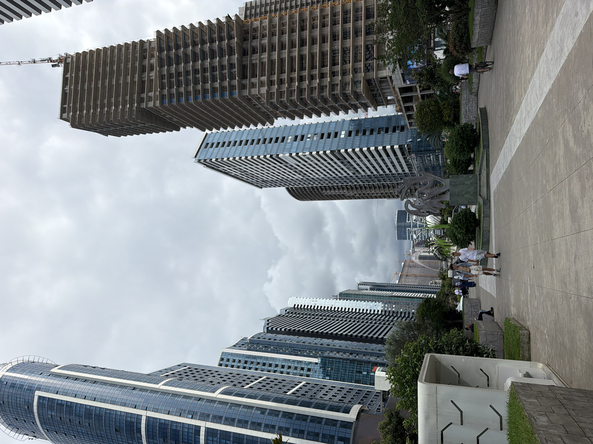 In Batumi, Georgia, a city promenade is depicted under a cloudy sky, featuring a row of modern high-rise buildings and one under construction. The foreground shows a paved walkway with a white stripe, lined by a planter with grass and a white, futuristic-looking structure with angled openings. Two young men are walking on the path, one wearing shorts and a t-shirt, the other in a white t-shirt and shorts. A man is sitting on a concrete bench in the background. To the left, a large, abstract sculpture stands on a pedestal. A person is riding a bicycle further down the path on the right side. The background is dominated by a cluster of skyscrapers, some fully constructed with glass facades, and one in the process of being built, showing its concrete floors and exposed framework, topped by a construction crane. More distant buildings and infrastructure, including a bridge and other cranes, are visible on the horizon.