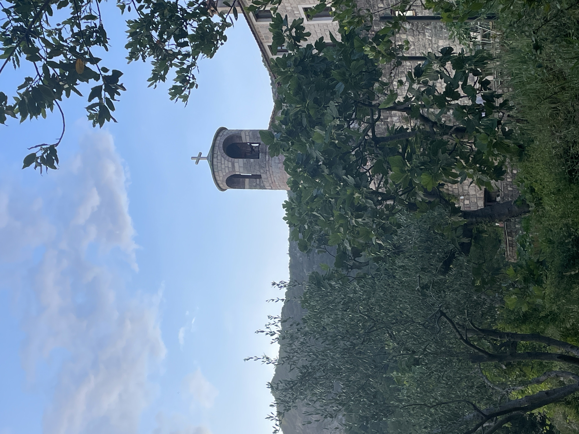 The image shows a low-angle view of a stone bell tower capped with a cross, likely part of a church, in Budva, Montenegro. The architecture is constructed from light-colored stone blocks and framed by lush green foliage of trees and bushes in the foreground. The sky is visible in the background, with blue areas and some white, soft clouds. The building has small rectangular windows. Part of a hill is also visible behind the structure.