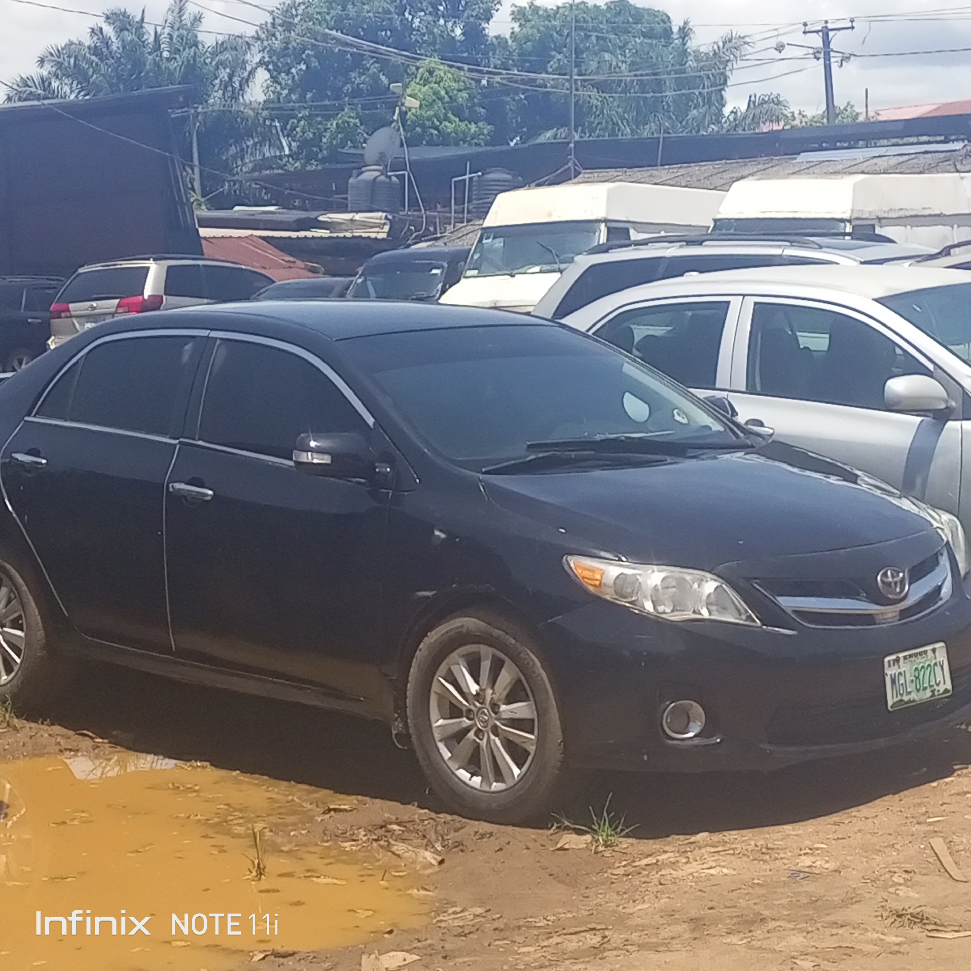 The image depicts a black Toyota Corolla parked in what appears to be a muddy lot in Fegge, Nigeria. Several other vehicles are visible in the background, including vans and cars. The environment appears to be a vehicle storage or sales area. Overheard power lines and vegetation, including palm trees, indicate an urban setting. A Nigeria license plate is visible on the front of the car. Text visible on the bottom corner identifies the photo was taken with an Infinix NOTE 11i. There is no visible interaction between humans and the objects described.