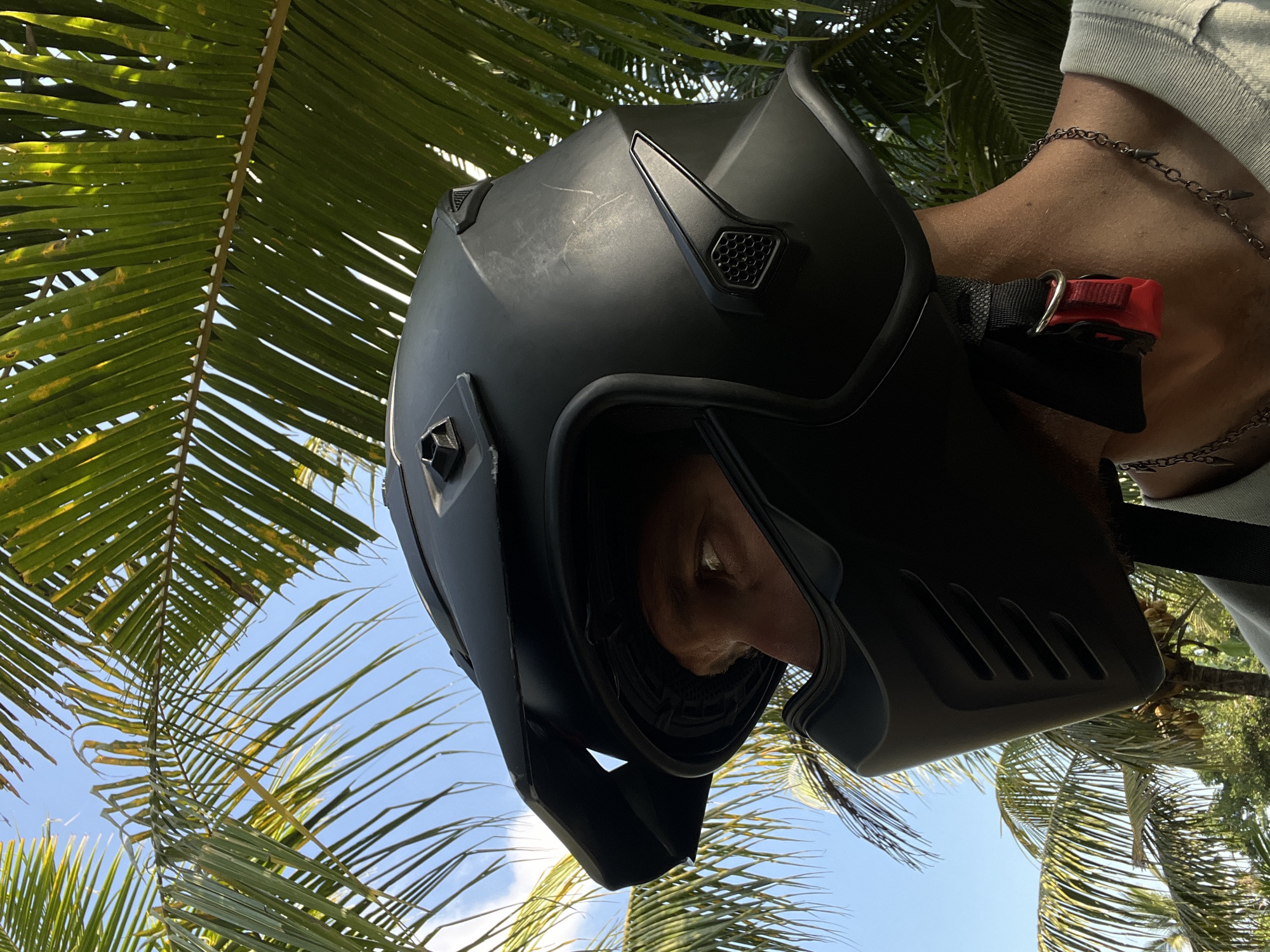 A person wearing a matte black motorcycle helmet with a protective face mask stands outdoors, partially obscured by palm fronds. The sky is blue with scattered clouds. The helmet has a prominent visor and several vents. The person's visible eye, a portion of their neck, and a chain necklace are also visible. The image appears to be taken at ground level, looking up. The location is specified as Kerambitan, Indonesia.