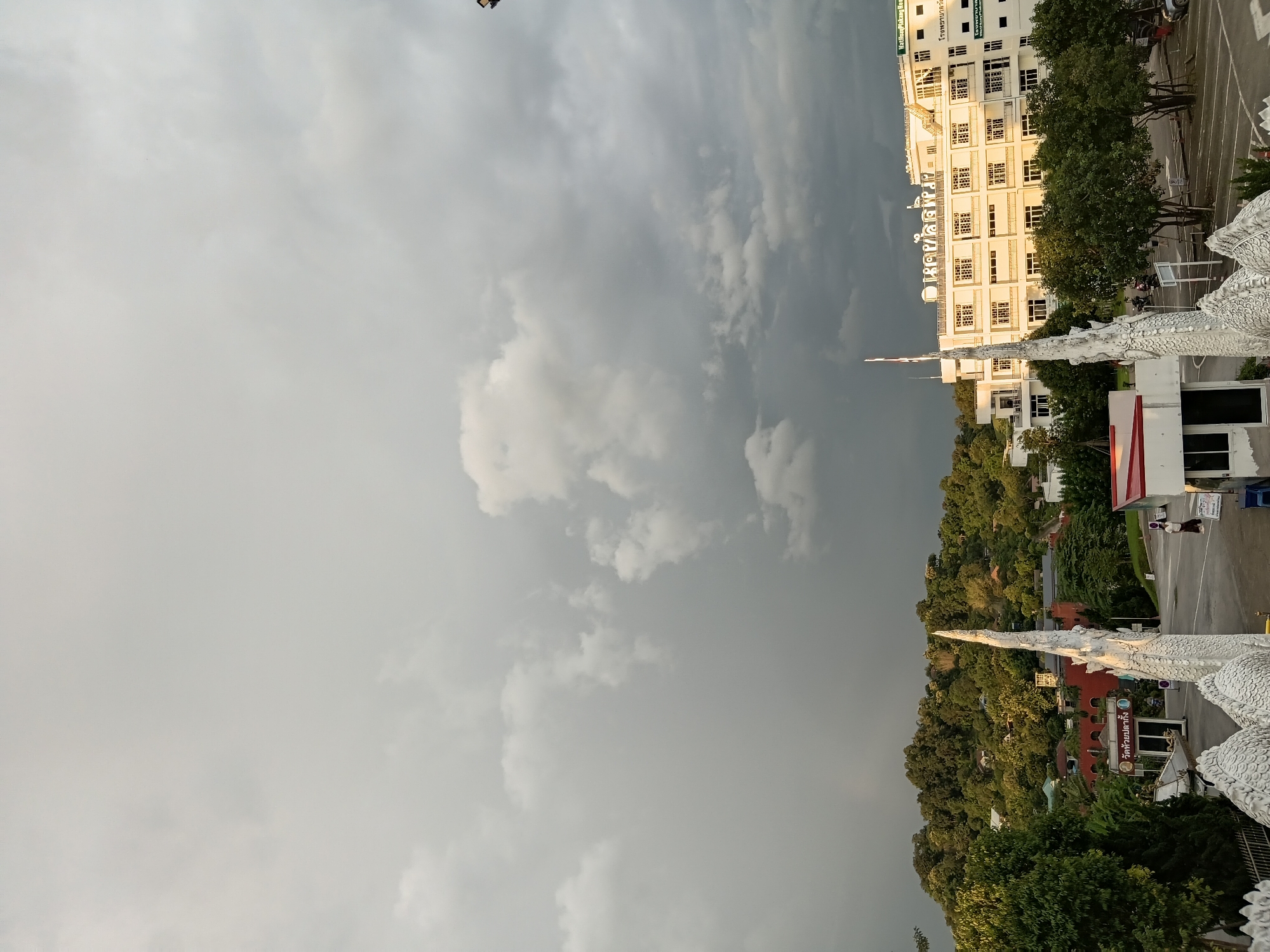 This is a medium shot, captured from an elevated perspective, showing a view of a street and buildings in the city of Ban Lao Pattana, Thailand. The sky is overcast with dark clouds, suggesting an impending storm. In the foreground, the white, ornate structure of a Naga, a mythical serpent, is visible. To the right, a modern, multi-story white building stands prominently, featuring numerous windows and balconies. Signs with Thai script are attached to the building. A smaller, red-roofed structure is situated to the left of the white building. A paved road with parking spaces is present in front of the buildings, and several vehicles are parked. Trees and a lush, green hillside with scattered houses form the background. One person, dressed in a white shirt and dark pants, is walking away from the camera on the right side of the scene. Another person, wearing a dark top and light pants, is walking towards the white building on the left. The overall impression is of a slightly ominous, yet potentially serene, urban landscape under a dramatic sky.