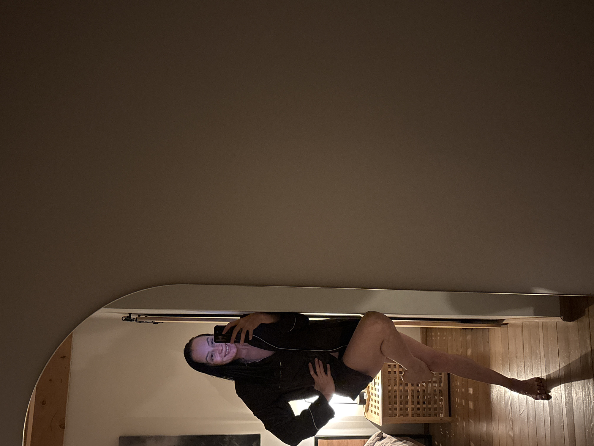 A woman is taking a selfie in a mirror, likely in a domestic interior. The room features wooden flooring and elements of neutral-toned decor. The woman is wearing dark sleepwear. It is not possible to determine the location of Chaikovsky, Russia, from the image.