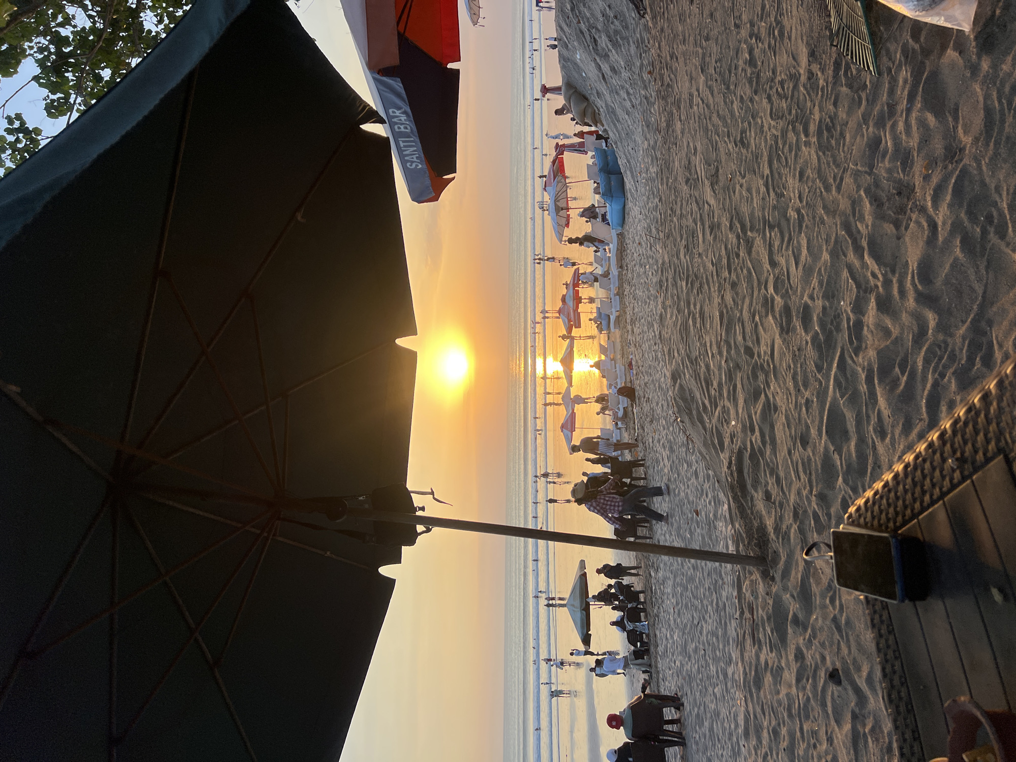 An exterior shot captures a beach scene in Kuta, Indonesia, at sunset. The foreground is dominated by a dark umbrella and a woven table with a smartphone on it. Beyond the table, the sandy beach is populated with numerous people, some seated on chairs, others standing or walking. Several beach umbrellas, including one with "SANTI BAR" written on it, dot the sand. In the middle ground, more individuals are visible on the wet sand near the ocean, and a few are wading in the water. The ocean stretches to the horizon, reflecting the warm orange glow of the setting sun. The sky is a soft gradient of orange and yellow, with the sun positioned prominently just above the horizon. Trees are visible in the upper right corner of the frame.