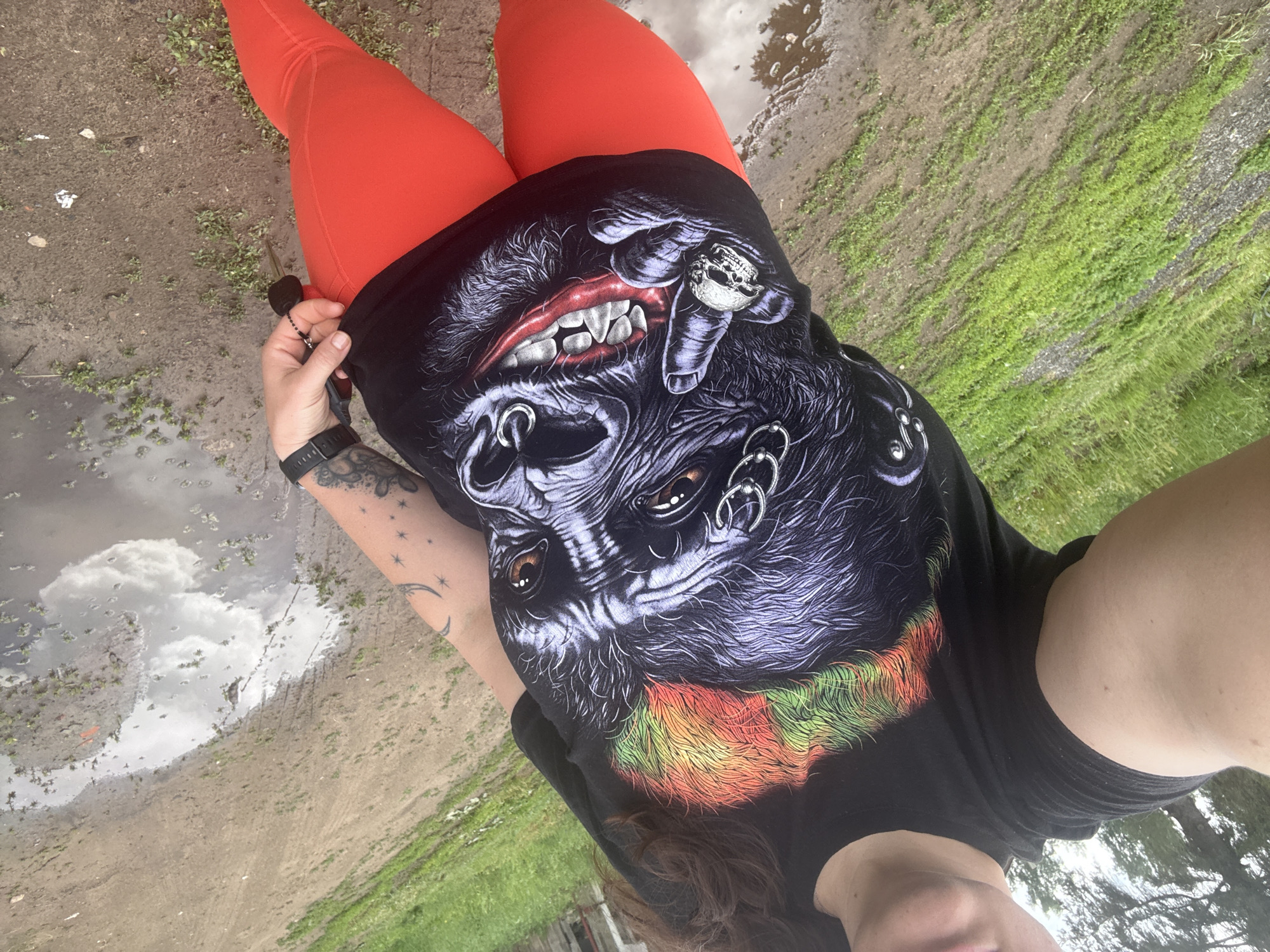 In Chaikovsky, Russia, a person is standing outside wearing a black t-shirt with a graphic of a gorilla's face wearing piercings and a skull ring on its finger, and bright orange leggings. The background consists of a dirt path with puddles of water and patches of green grass, suggesting an outdoor, possibly rural or park-like, setting. The person is holding a set of keys in their right hand.