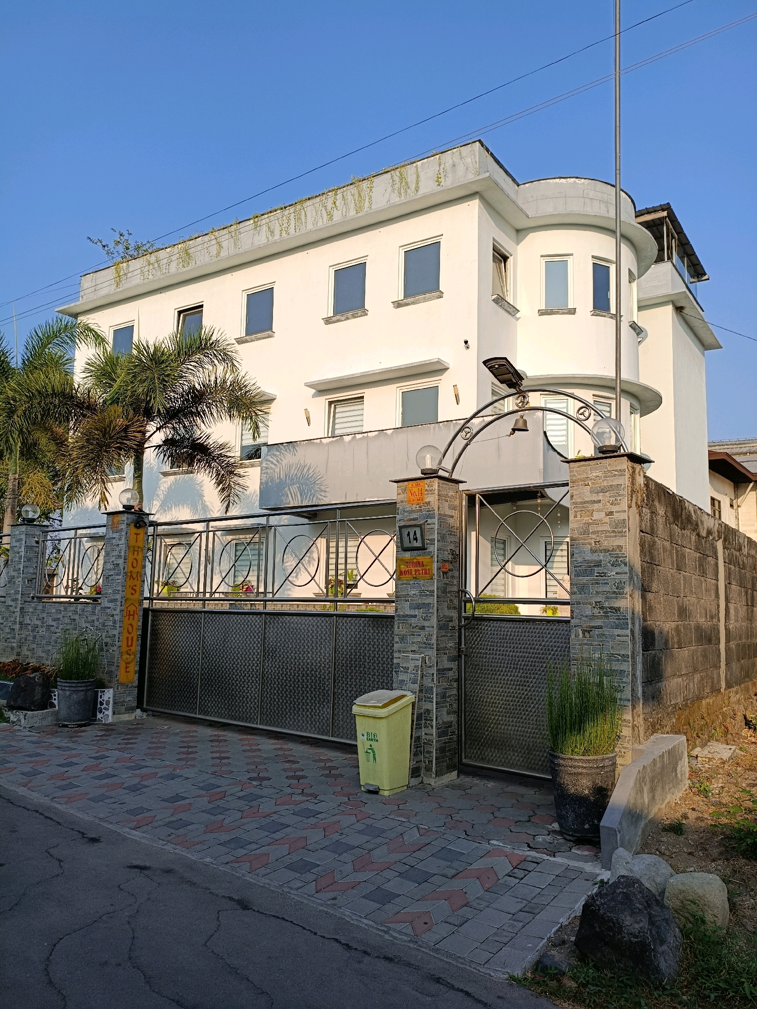 The image shows a modern, multi-story white building with a curved facade on the right side. The building has several rectangular windows and a balcony. In front of the building, there is a metal gate with decorative circular patterns and a sign that reads "THOM'S HOUSE". The gate is flanked by stone pillars and a stone wall. Palm trees are visible to the left of the building. A yellow trash can and potted plants are placed near the gate. The ground is paved with interlocking bricks. The scene appears to be on a street in a residential or commercial area, possibly in Maguwoharjo, Indonesia, given the architectural style and vegetation. The sky is clear and blue, suggesting it is daytime. There are no people visible in the image.
