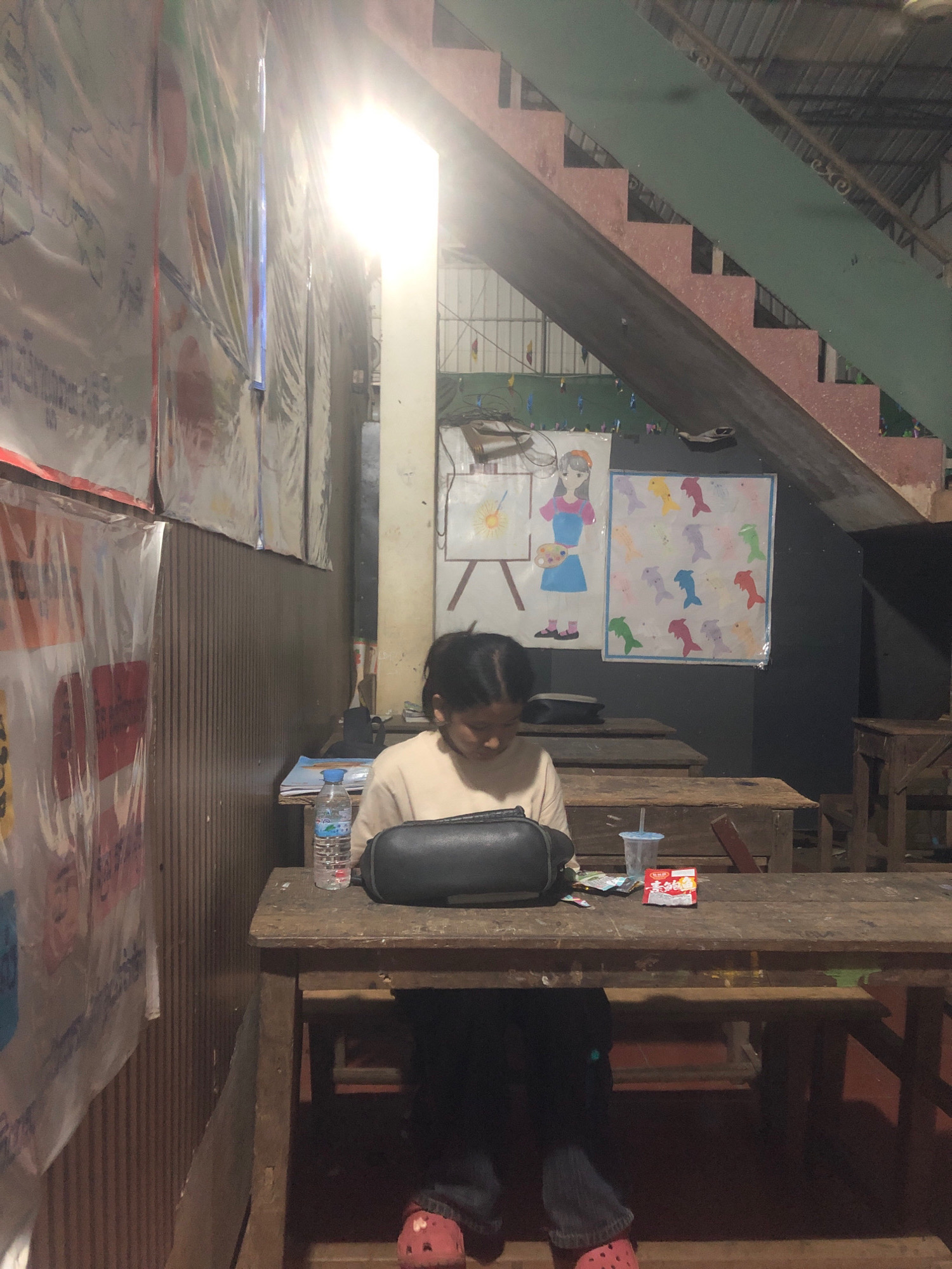 In Kanhchriech, Cambodia, a young person with dark hair sits at a wooden desk, engrossed in reading a book. They are wearing a light-colored long-sleeved shirt and dark pants, with bright pink clogs visible on their feet. The desk is cluttered with a water bottle, a plastic cup with a straw, a small packet of snacks, and other assorted items. Behind the person, posters depicting various images, including a drawing of a girl painting and a collage of colorful fish, are displayed on the wall. A set of wooden stairs with red steps and a green railing is prominent in the background, leading upwards. The overall environment appears to be a dimly lit indoor space, possibly a classroom or a community center.