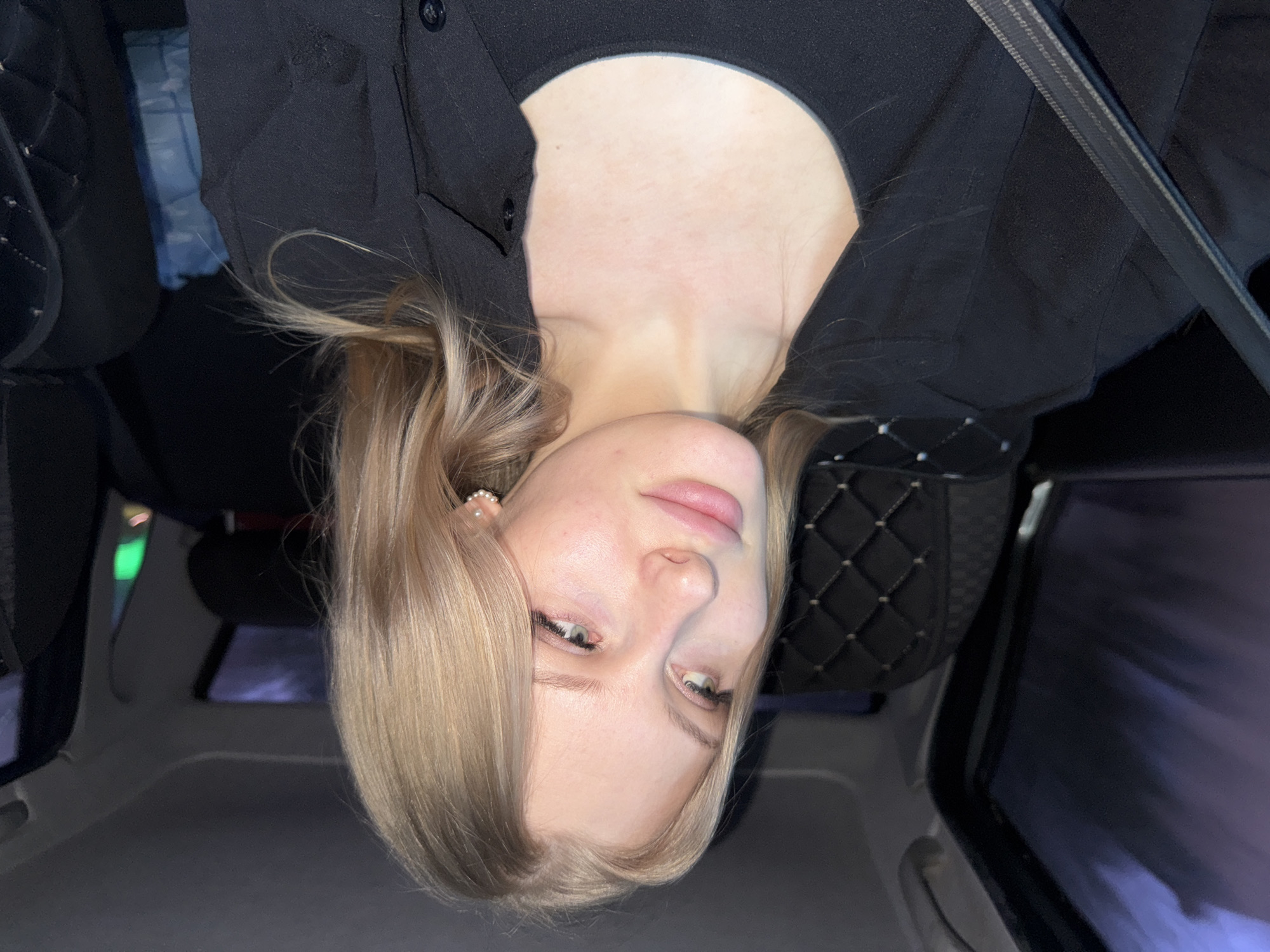 A fair-skinned young woman with blonde hair is positioned inside a vehicle, possibly a car, with her head tilted downward. She is wearing a black jacket over a dark top. The interior of the vehicle features a black seat cover with a quilted pattern, and a safety belt strap is visible. The scene suggests an urban environment, given the vehicle context, and the presence of a reflection of city lights on the car window. The precise location is not clear, and there is no verifiable evidence suggesting the location is Perm, Russia.