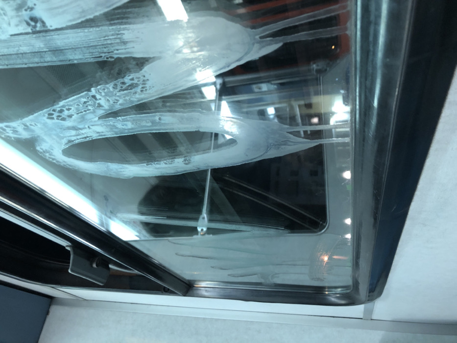 The image displays an upward-angled view through a clear glass surface, likely a window or skylight, significantly covered in dried white residue. The residue forms distinct patterns, including circular shapes, streaky lines, and bubbly clusters, suggesting dried soap suds, hard water stains, or an improperly cleaned surface.
The glass is encased in a dark, possibly metallic or plastic, frame visible around the edges. In the lower-left section of the frame, a black handle or latch mechanism is evident. Through the relatively clearer parts of the glass, reflections or blurred views of an interior space are visible, showing lighter-colored surfaces and darker, indistinct structural elements in the background. The lighting appears bright and natural, indicating daytime.
No people or specific activities are depicted. The scene suggests an interior environment, possibly within a vehicle like a camper van or bus, or a room with a skylight. The photograph provides no specific visual cues to confirm the external location of Kallithea, Greece. There is no visible text.