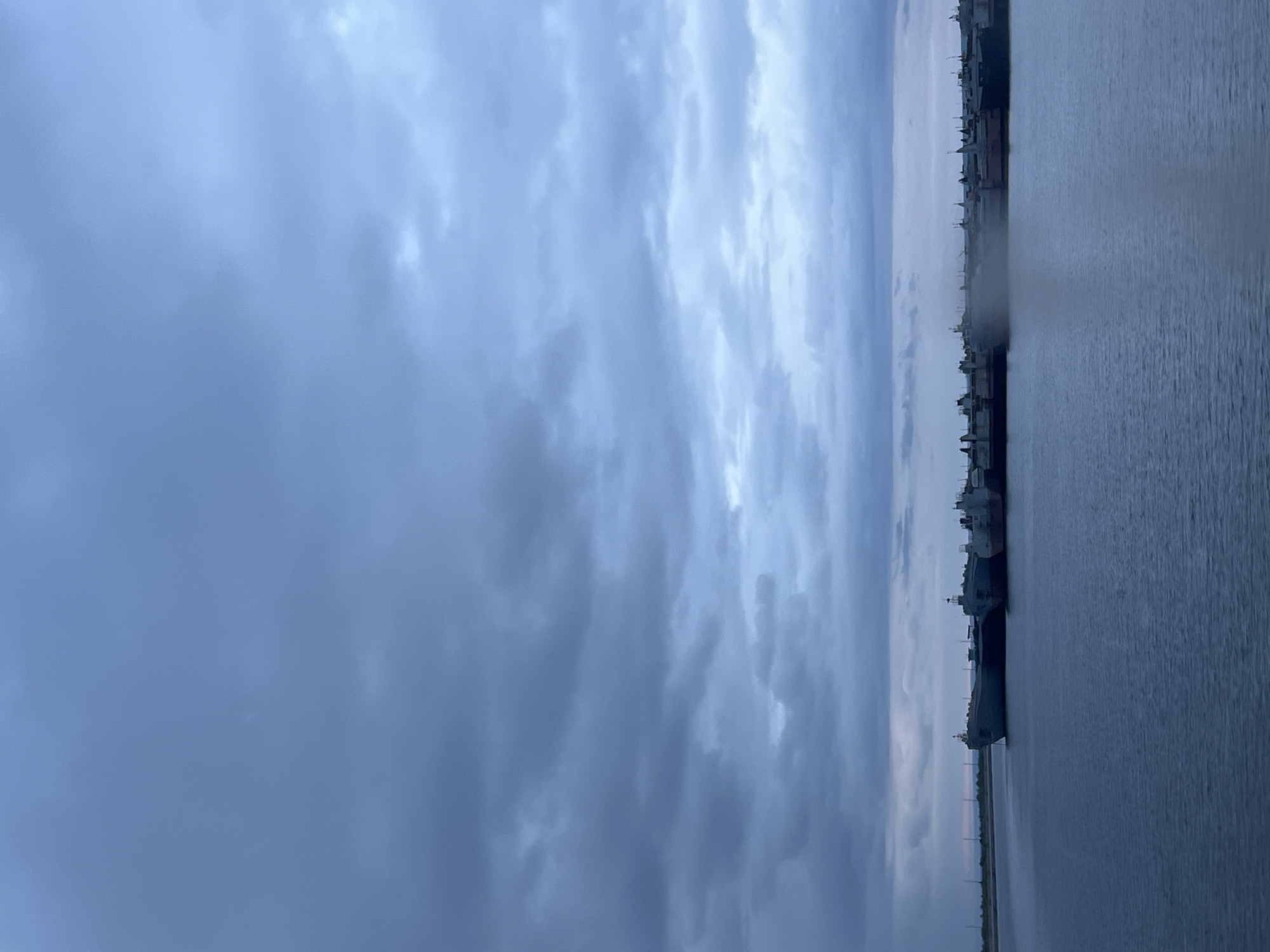 The image displays an exterior, wide-angle view of a large body of water, identified as a section of the Kama River in Chaikovsky, Russia. The scene is captured under heavily overcast skies, suggesting either twilight conditions or a fully clouded day, imparting a cool, dim, blue-grey illumination across the entire landscape.
The upper two-thirds of the frame are occupied by a dense canopy of stratocumulus clouds, varying in tone from lighter blue-grey in the upper central and right portions to darker, more saturated grey-blue near the horizon and in the lower cloud formations. The cloud cover is extensive and appears uniform, with no visible breaks to direct sunlight.
The lower third of the image depicts the still surface of the water, which is a consistent dark blue-grey, mirroring the colors of the sky. The water's surface is smooth with only minimal, subtle ripples, indicating calm conditions.
Along the distant horizon, a low-lying landmass serves as a bank, stretching horizontally across the entire width of the image. This bank is densely lined with a multitude of large vessels, including what appear to be cargo ships or freighters, moored closely together. These vessels are predominantly dark in silhouette against the slightly lighter horizon and sky, with the leftmost ship exhibiting a distinct grey hull. Their forms suggest substantial industrial vessels, featuring visible superstructures, masts, and deck equipment. In the central-right area, directly above the water and in front of a cluster of these vessels, a localized area of atmospheric mist or vapor is discernible. To the far left, a faint, light pinkish-blue atmospheric glow is visible just above the distant land strip.
There are no visible human figures, specific actions, or immediate foreground objects in the image. The scene conveys a quiet, expansive industrial waterfront environment.
