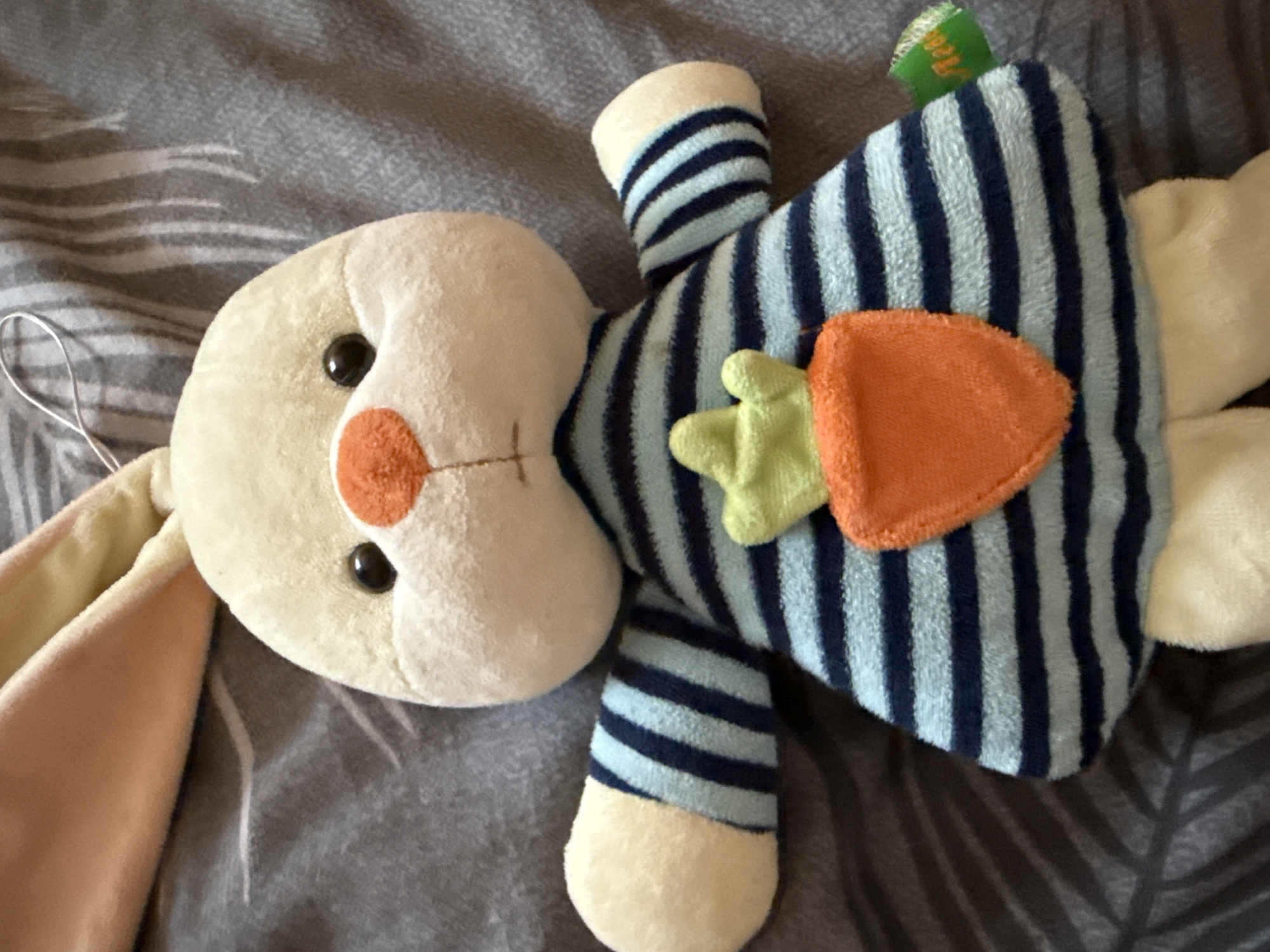 This is a close-up, eye-level shot of a plush toy rabbit lying on a grey, textured surface that appears to be bedding. The rabbit is light cream-colored with a bright orange nose and black button eyes. It is wearing a blue and navy striped shirt with a green leafy carrot applique on the front. The rabbit's ears are light pink on the inside and cream on the outside. There is a thin white string attached to the top of the rabbit's head. A small green tag with illegible text is attached to the rabbit's right side. The background is a blurred grey fabric with a subtle pattern. There is no indication of the city of Okhtyrka, Ukraine, in this image.