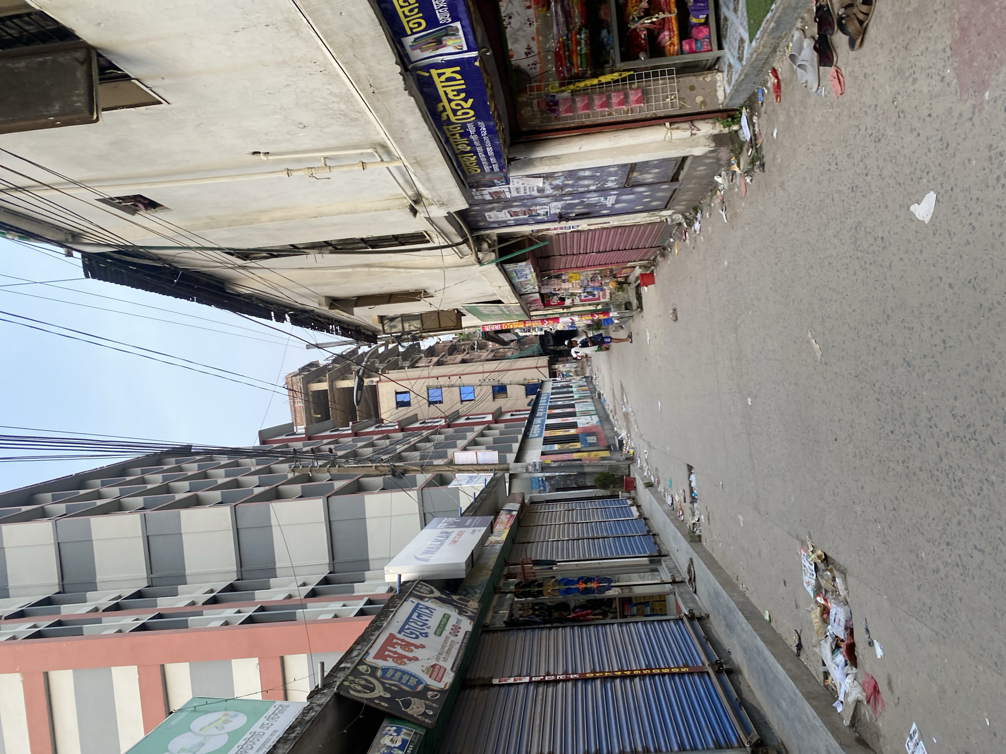 The image depicts a street scene in Dhaka, Bangladesh. The perspective is angled, looking down a street lined with buildings and shops. Overhead power lines are visible against the sky. The shops are identifiable by their signage, some in Bengali script. The street surface is marked with litter and debris. People are present in the distance.