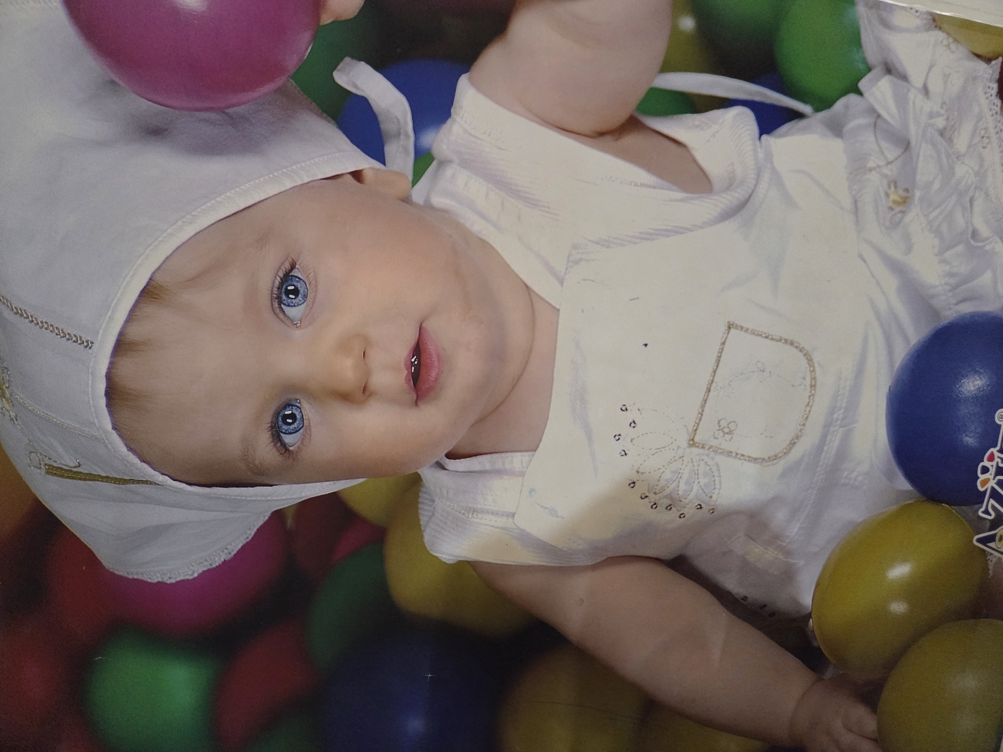 A baby with blue eyes and a white hat is lying amidst colorful plastic balls. The baby is holding a purple ball. The baby is wearing a white outfit with embroidery. The background consists of a variety of colored plastic balls, including pink, green, yellow, and blue. The location is unknown and there is no indication the photo was taken in the city of Pleven, Bulgaria.