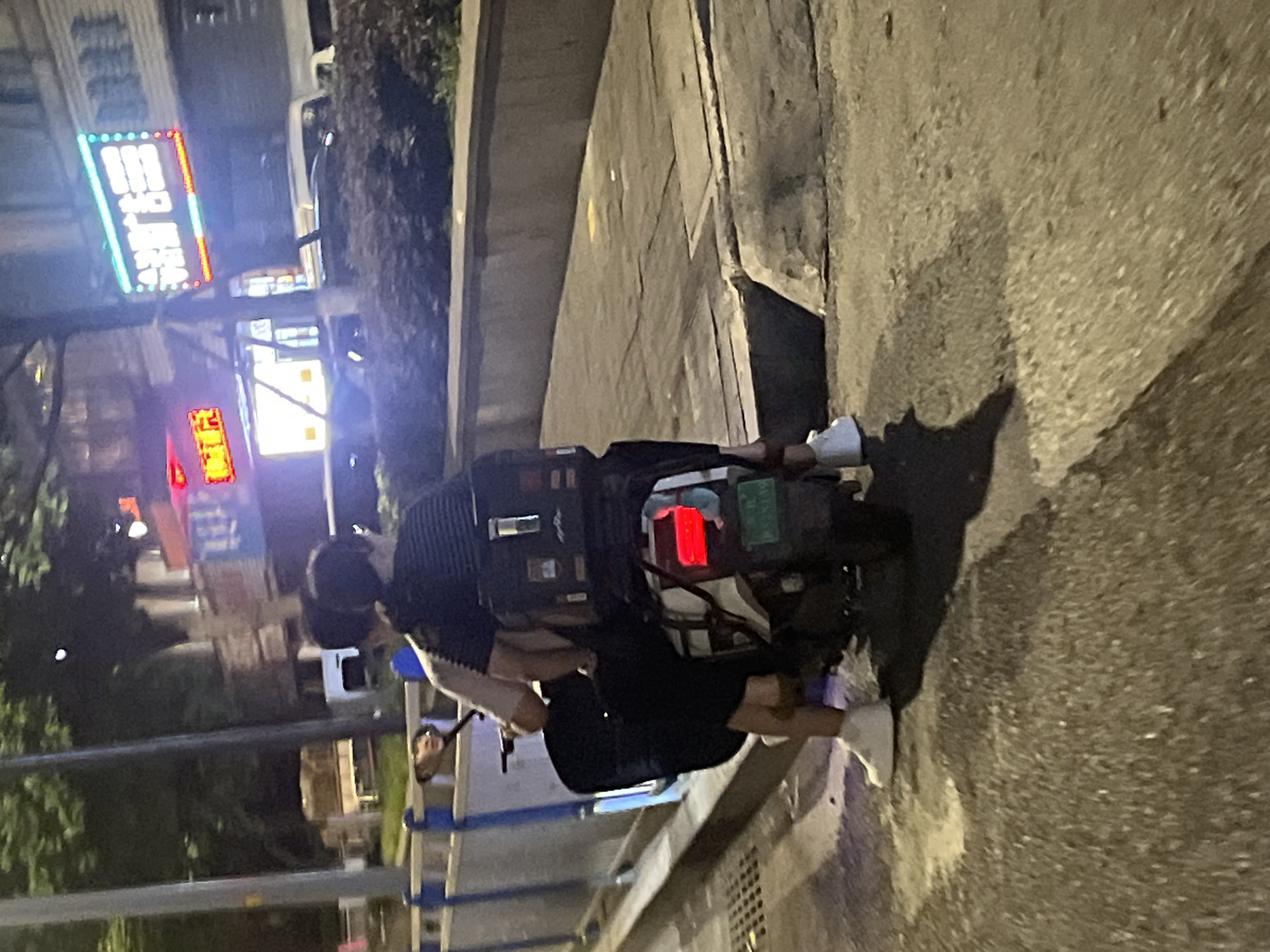 This image depicts a nocturnal urban scene in Jiulongpo District, Chongqing, China. Two individuals are positioned on a white electric scooter, facing away from the viewer. The person in the driver's seat wears a light-colored short-sleeved shirt and dark shorts, with white athletic shoes visible on their feet. Their reflection, looking towards the left, is discernible in the scooter's right rearview mirror. The second individual, seated behind the driver, wears a dark, horizontally striped short-sleeved shirt. A large, rectangular black delivery box is secured to the rear of the scooter, behind the passenger.
The scooter's rear brake light is illuminated red, and a green Chinese license plate, bearing the characters "渝A 7M139," is affixed below it. The scooter is situated on a dark paved surface. To the left, a grey metal barrier runs parallel to the scooter. To the right, a raised concrete curb separates the paved area from a sidewalk composed of concrete slabs. Beyond the sidewalk, a low concrete wall fronts a strip of dark foliage.
In the background, multiple buildings are visible under artificial illumination. Several bright neon signs are present; one prominent red and yellow sign displays Chinese characters, and another large, rectangular LED sign, with white characters on a blue and red background, is also visible. A white vehicle, possibly a van, is partially discernible in the distance behind the foliage. Streetlights and building lights contribute to the ambient illumination of the scene.