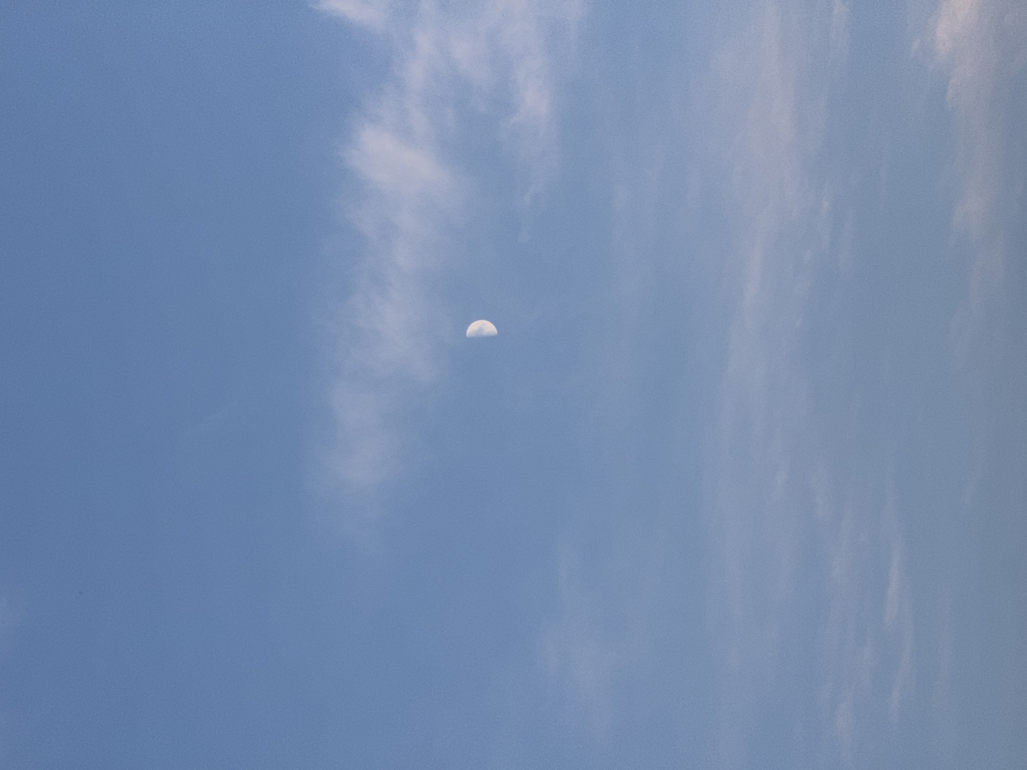 This image captures a daytime sky with wispy clouds and a crescent moon. The sky is a clear, pale blue, typical of a fair weather day. The moon, illuminated by the sun, is visible as a sliver in the upper center of the frame, suggesting it is either early morning or late afternoon. The clouds are thin and scattered, allowing ample sunlight to filter through. The overall impression is serene and tranquil, with the vastness of the sky dominating the scene. No people, objects, text, or specific location cues are discernible, beyond the implied open sky.