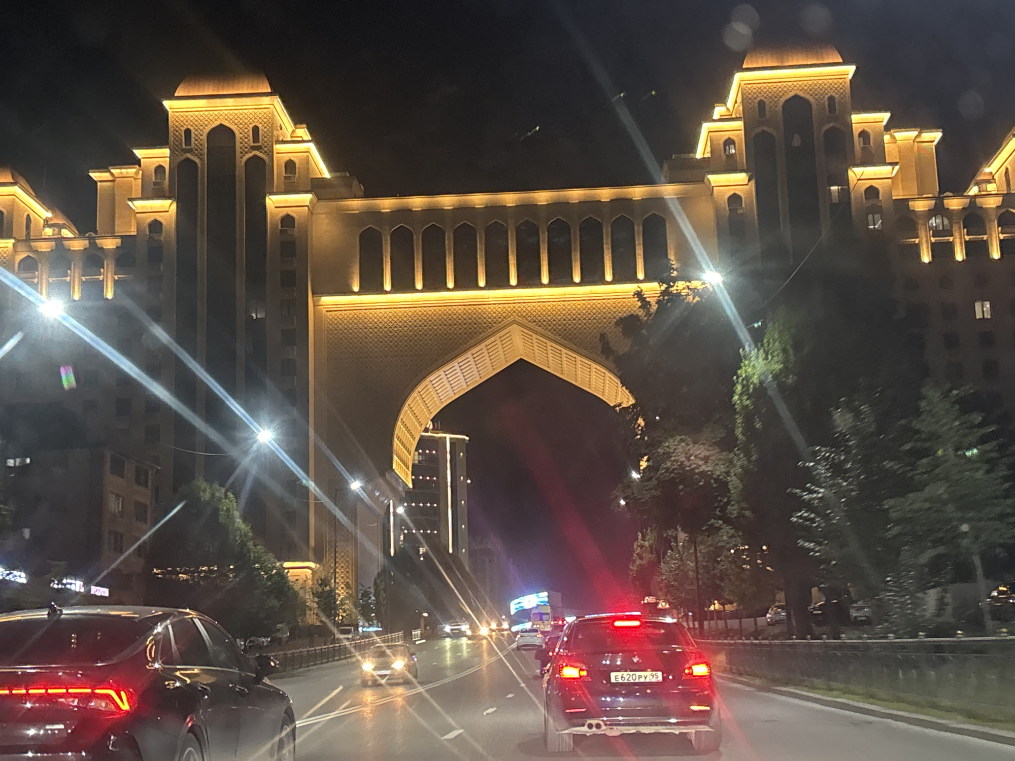 The image captures a nighttime view of a city street, likely in Grozny, Russia, featuring a large, illuminated building with Islamic architectural elements. The building is ornately decorated with golden lights highlighting its arches, towers, and patterned facades. In the foreground, several cars are visible on a multi-lane road. The car in the immediate foreground on the right is a dark-colored sedan with its brake lights illuminated. The car in the foreground on the left is also a dark sedan, partially visible. The road is lit by streetlights and the building's illumination. Trees line the side of the road, and other buildings are visible in the background. The overall scene suggests a well-lit urban environment at night.