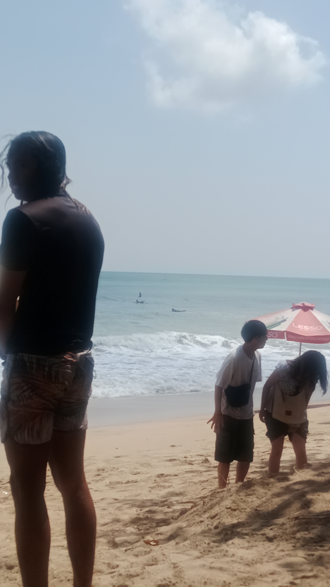 At the sandy beach of Kuta, Indonesia, under a bright, hazy sky with scattered clouds, a person stands in the foreground with their back to the camera. They are wearing a dark t-shirt and patterned shorts. Further down the beach, two individuals are bent over, appearing to interact with the sand. One is wearing a light-colored t-shirt and dark shorts, with a dark bag slung across their body. The other person, to their right, is wearing a light-colored top and dark shorts, also with a bag. In the background, the turquoise ocean meets the shore with gentle waves, and several surfers can be seen in the water. A red and white striped beach umbrella with the word "LESSO" is visible near the two bent-over individuals. The sand is textured with footprints and shadows cast by an unseen object.