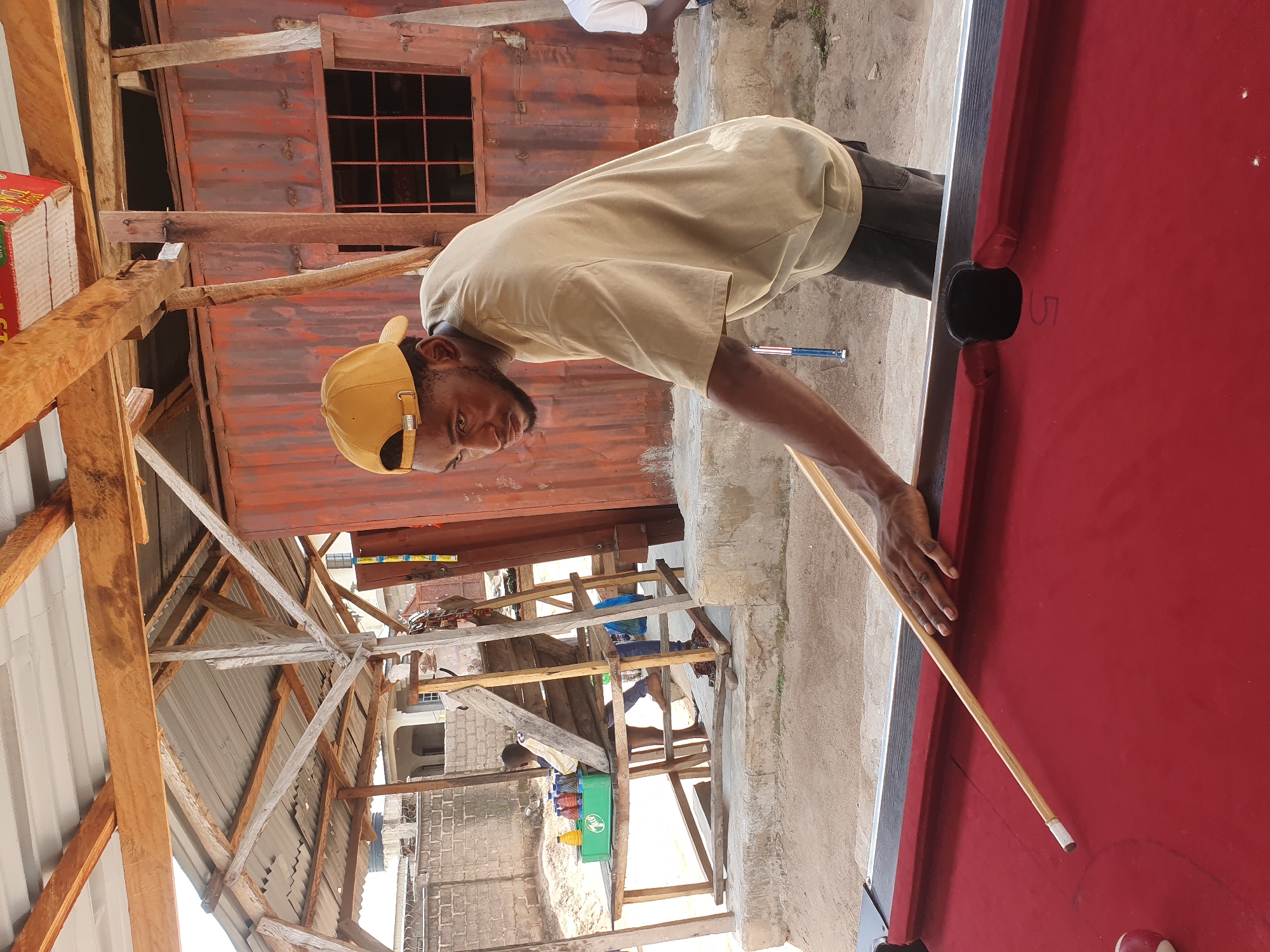 In Lokoja, Nigeria, a young Black male wearing a tan t-shirt and a yellow baseball cap is playing pool. He is standing on a dirt ground next to a red pool table and is in the process of taking a shot. The pool table is set up outdoors under a rustic, open-air shelter made of corrugated metal roofing and wooden beams. In the background, there is a rusty corrugated metal building with a window, and to the left, a wooden structure with a green container holding bottles. Another person is visible sitting behind this wooden structure. The scene suggests a casual, informal setting for playing pool.