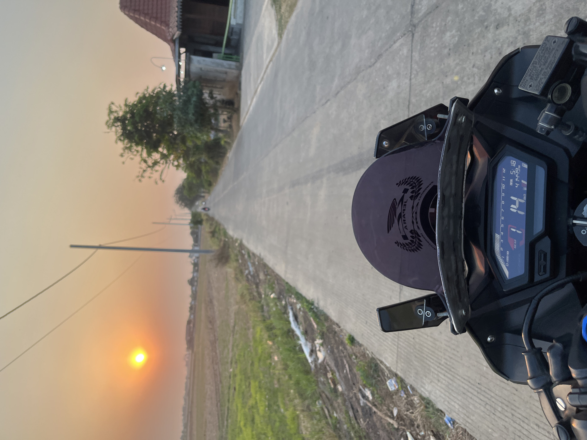The image depicts a first-person perspective from a motorcycle traveling down a paved road in Pecangaan, Indonesia, during sunset. The sun, a bright orange orb, is visible in the upper left quadrant, casting a warm glow over the scene. To the left of the road, a field of green and brown vegetation stretches towards the horizon, with scattered debris visible along the roadside. To the right, trees and a traditional Indonesian house with a red tiled roof are present. The road itself is a light gray asphalt with visible cracks and some debris along the edges. In the foreground, the motorcycle's dashboard displays a speed of 14 km/h and a gear indicator showing "1". The handlebars and part of the motorcycle's front fairing are also visible. A few distant figures on motorcycles can be seen further down the road. The sky is a soft gradient of orange and pale yellow, indicating the end of the day. Electrical wires and poles are present along the side of the road.