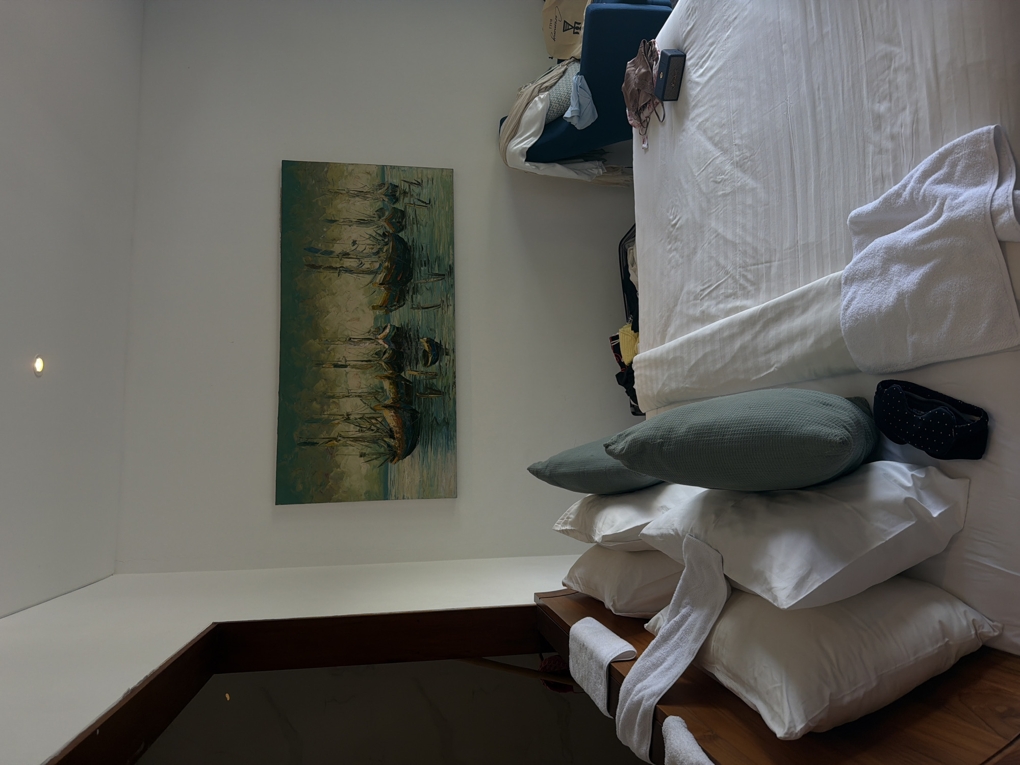 The image shows the interior of a room, likely a hotel room, with a bed dominating the scene. The bed is covered with white linens and a white towel is folded on top. There is a stack of pillows on the side of the bed, including a larger green pillow. A black sleep mask rests atop the pillows. To the left of the bed, a painting of ships at sea hangs on the wall. In the background, a blue chair and additional luggage can be seen. The location is identified as Petulu, Indonesia. There are no people visible in the image.