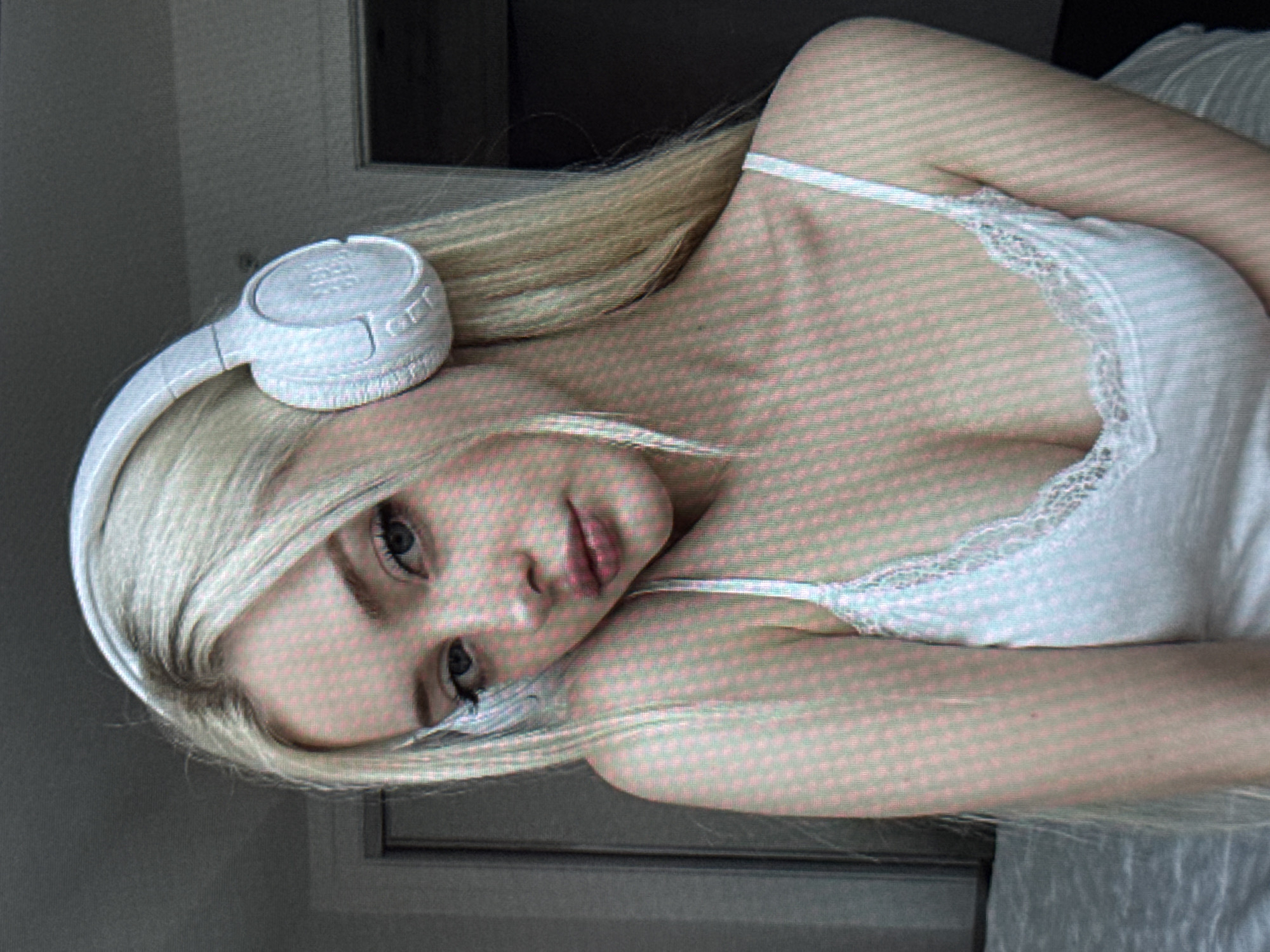 A head-and-shoulders portrait features a young woman with long, straight blonde hair, fair skin, and blue eyes. She is captured with her head tilted, looking directly at the camera with a soft, neutral expression. She wears white over-ear headphones, with the "JBL" logo clearly visible on the right earcup. Her blonde hair cascades around her shoulders, partially obscuring the left headphone. She is dressed in a light-colored camisole or tank top, possibly white, which features delicate lace detailing along the neckline. A thin spaghetti strap is visible over her right shoulder. The background is softly blurred, suggesting an indoor setting with a light-colored wall and some darker elements that could be furniture or a window frame. The overall image has a slightly grainy appearance. The woman's pose conveys a sense of relaxation, possibly reclining. No specific visual cues within the image indicate the location is Varna, Bulgaria.