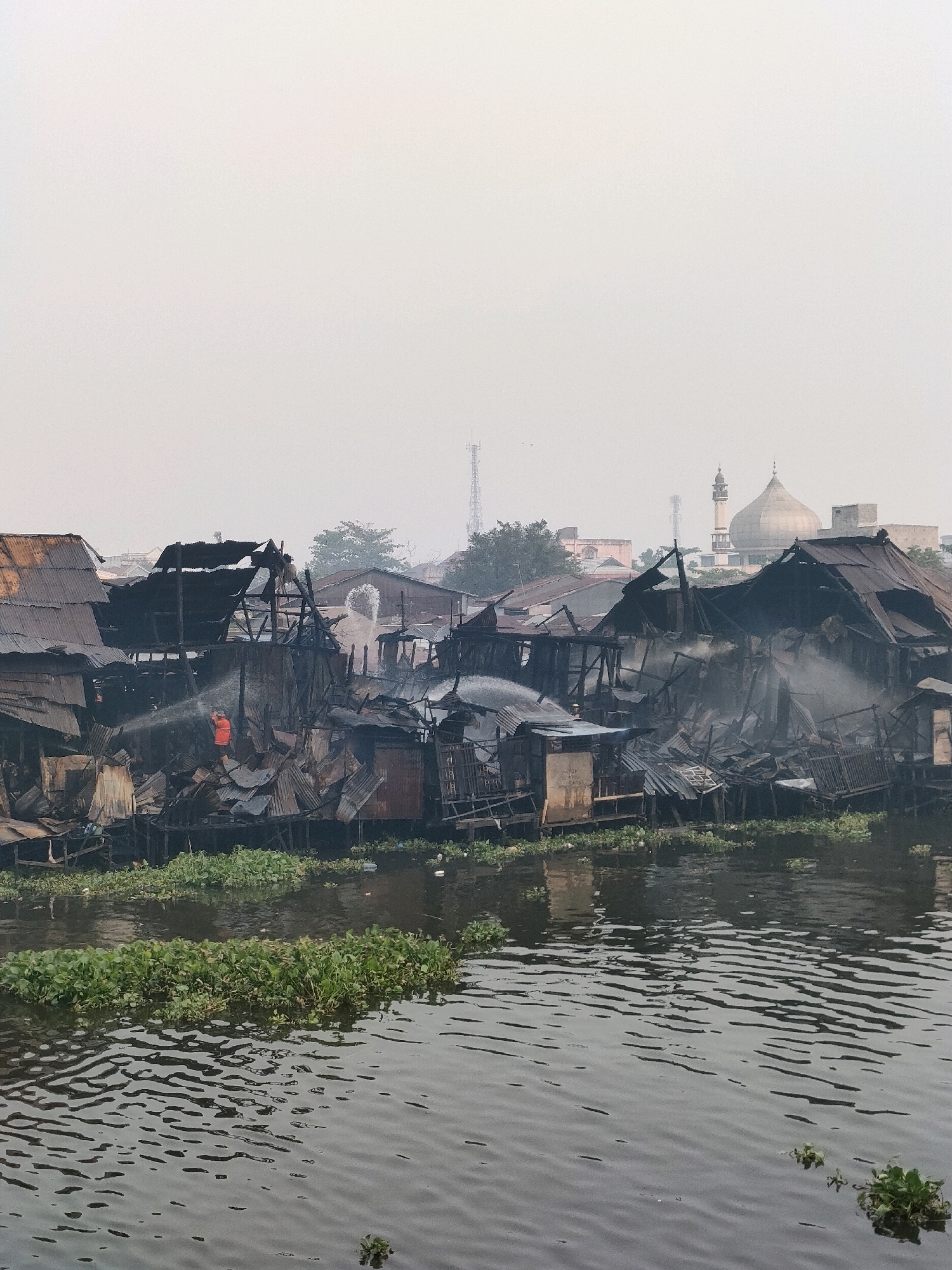 The image depicts a devastating fire in a riverside slum, likely located in Banjarmasin, Indonesia, given the architecture and the presence of a mosque in the background. The scene is characterized by damaged, partially burned structures built on stilts over a murky body of water. Smoke billows from the wreckage of tin and wooden houses, some of which have collapsed. In the foreground, a person in an orange vest, presumably a firefighter, is seen spraying water on the fire with a hose. Water hyacinths float on the surface of the river, interspersed with debris from the fire. The sky is overcast and hazy, contributing to the somber atmosphere. In the distance, a prominent mosque with a dome and minaret stands as a stark contrast to the destruction in the foreground, and communication towers are visible. The overall impression is one of a community reeling from a significant disaster.