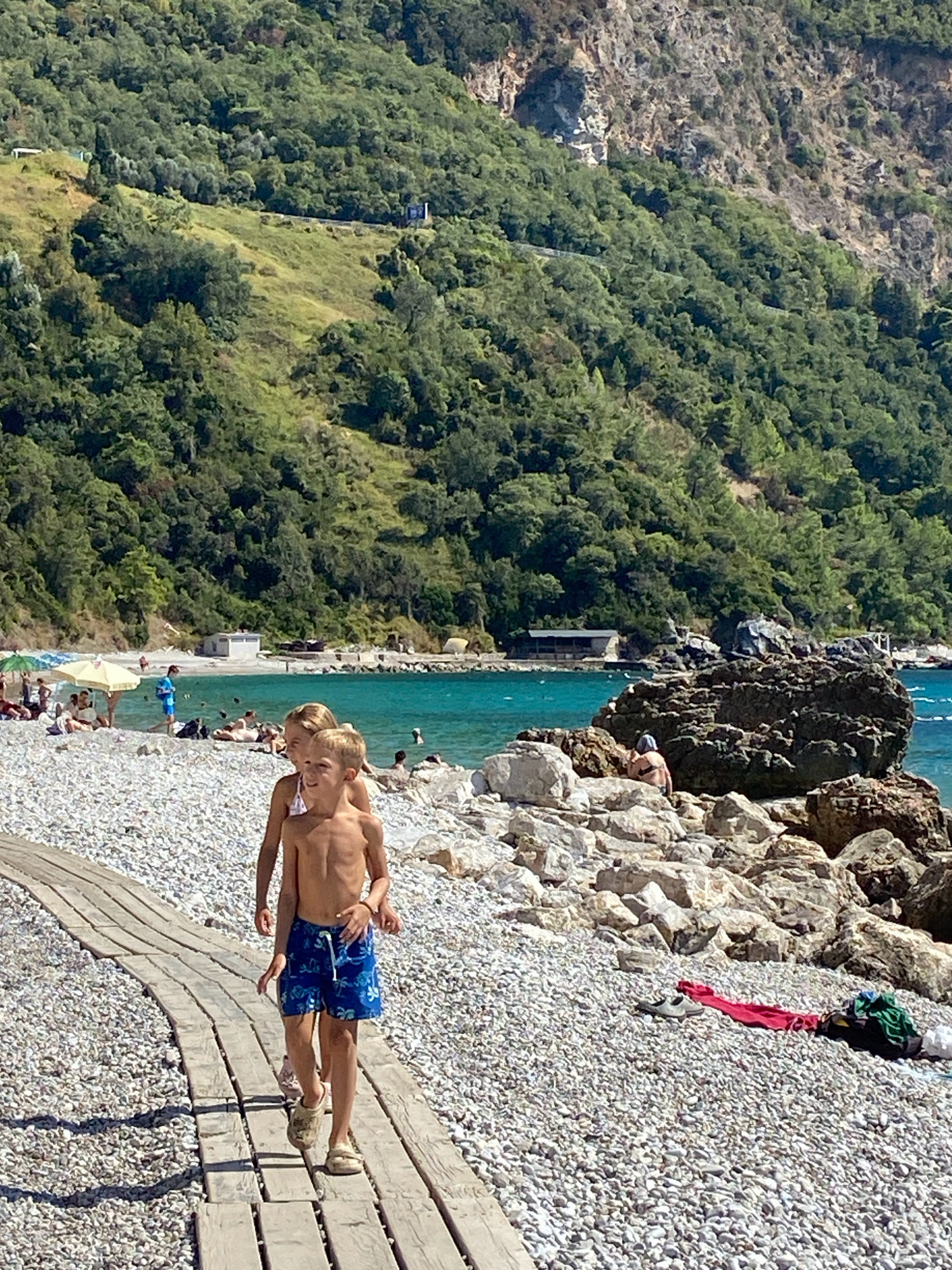 The image depicts a sunlit, pebble beach in Prijevor, Montenegro. In the foreground, two children, a boy and a girl, walk on a wooden boardwalk. The boy, shirtless, wears blue patterned swim trunks, and the girl, partially visible, wears a bikini top. The beach is covered in white and grey pebbles, with a rocky outcrop to the right. Beyond the beach, the turquoise Adriatic Sea is visible, with several people lounging on the shore and swimming. In the background, a densely forested, steep hillside rises, with a glimpse of a road or path winding along its contours. A white building, possibly a restroom facility, is situated near the water's edge. The scene is characteristic of a popular coastal tourist destination.